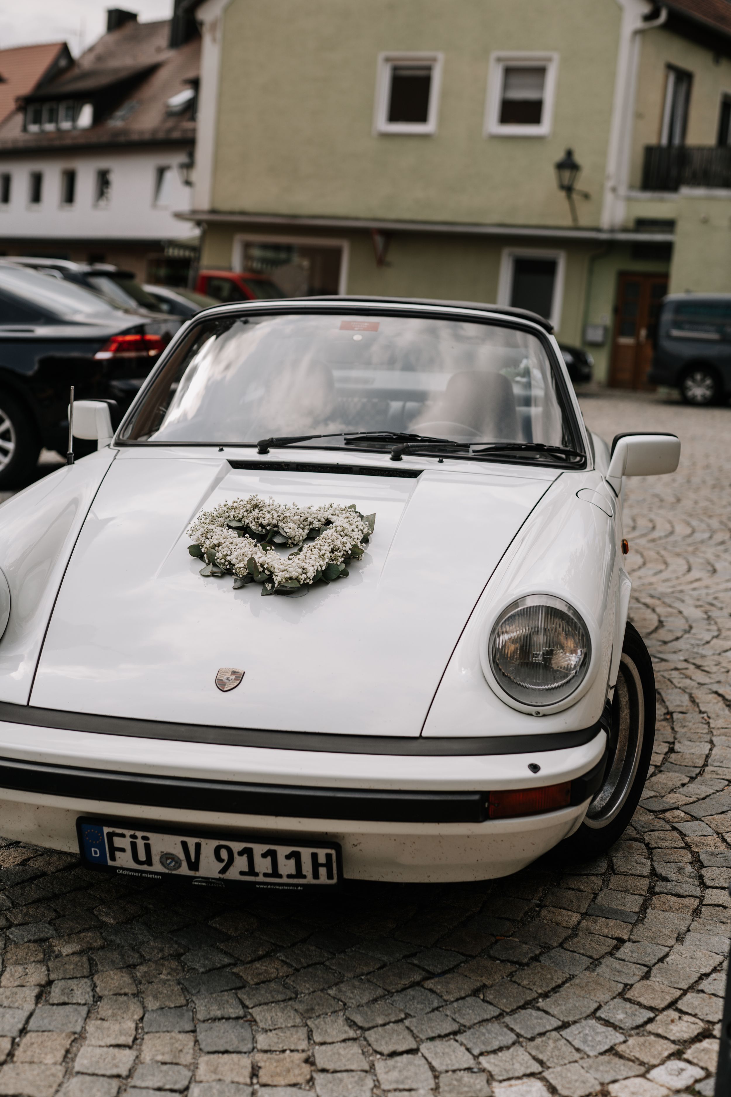 A classic white Porsche decorated with a floral wreath sits on cobblestone streets.