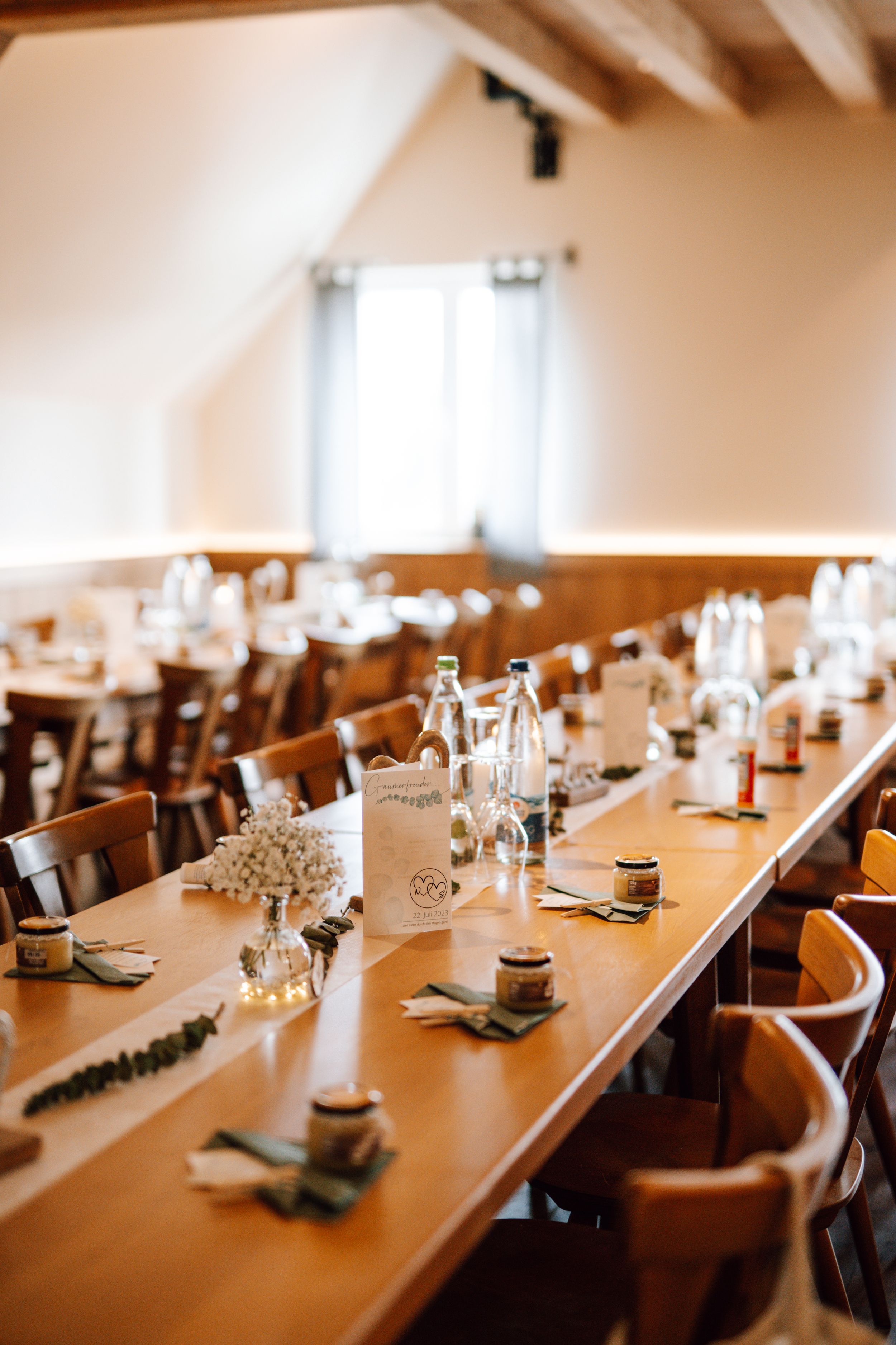 Long banquet tables set for wedding reception in bright room with exposed beams.