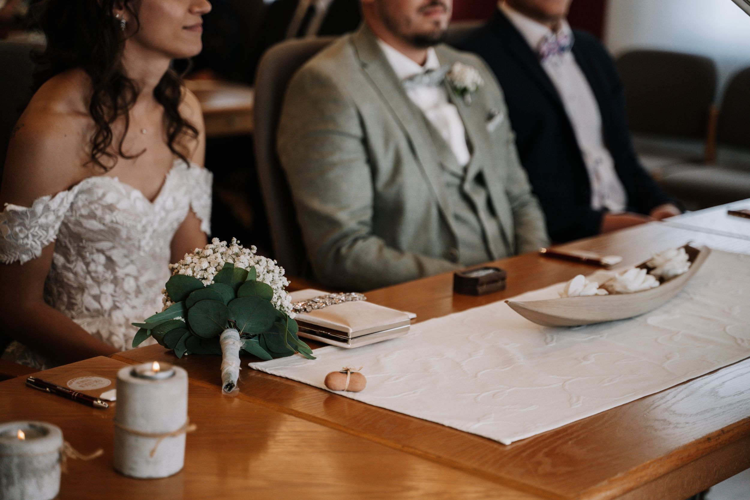 A couple sits at a wooden table during their wedding ceremony with a small floral arrangement and paperwork.
