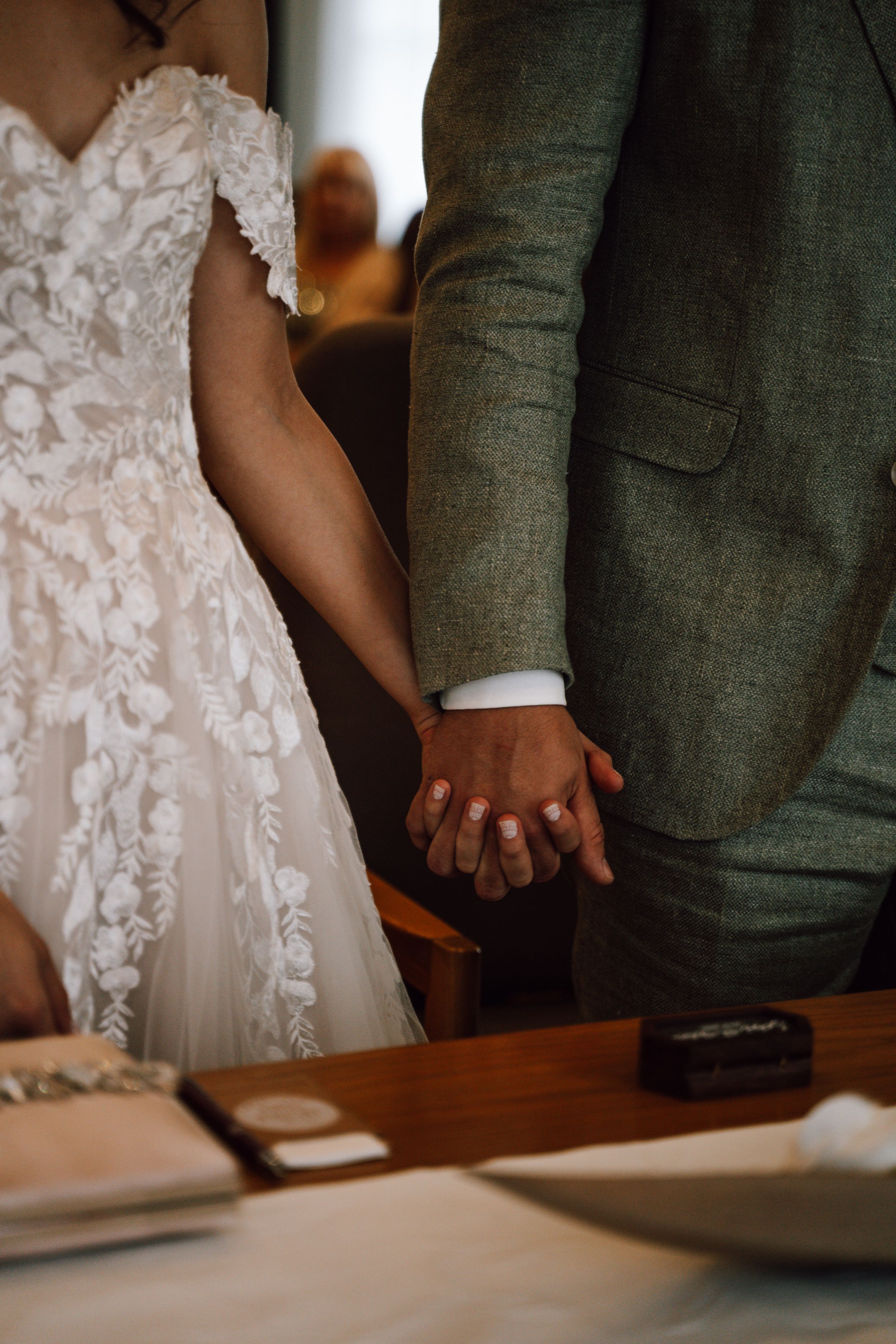 Close-up of clasped hands showing lace wedding dress and sage green suit details.