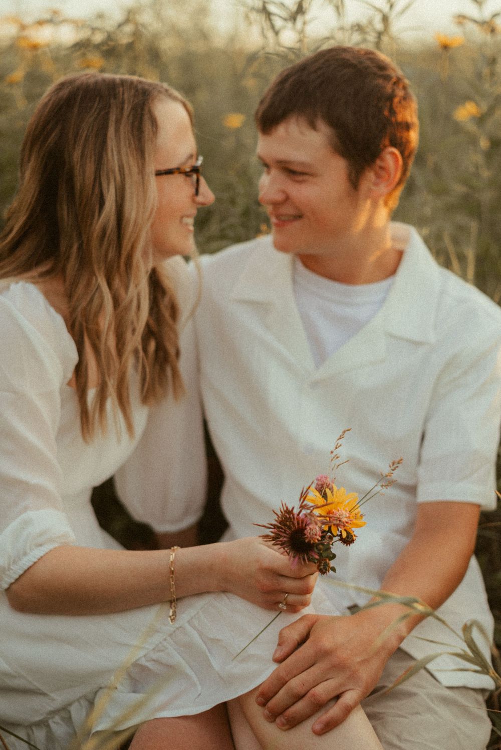 Couple sharing intimate moment in sunlit field with small wildflower bouquet.