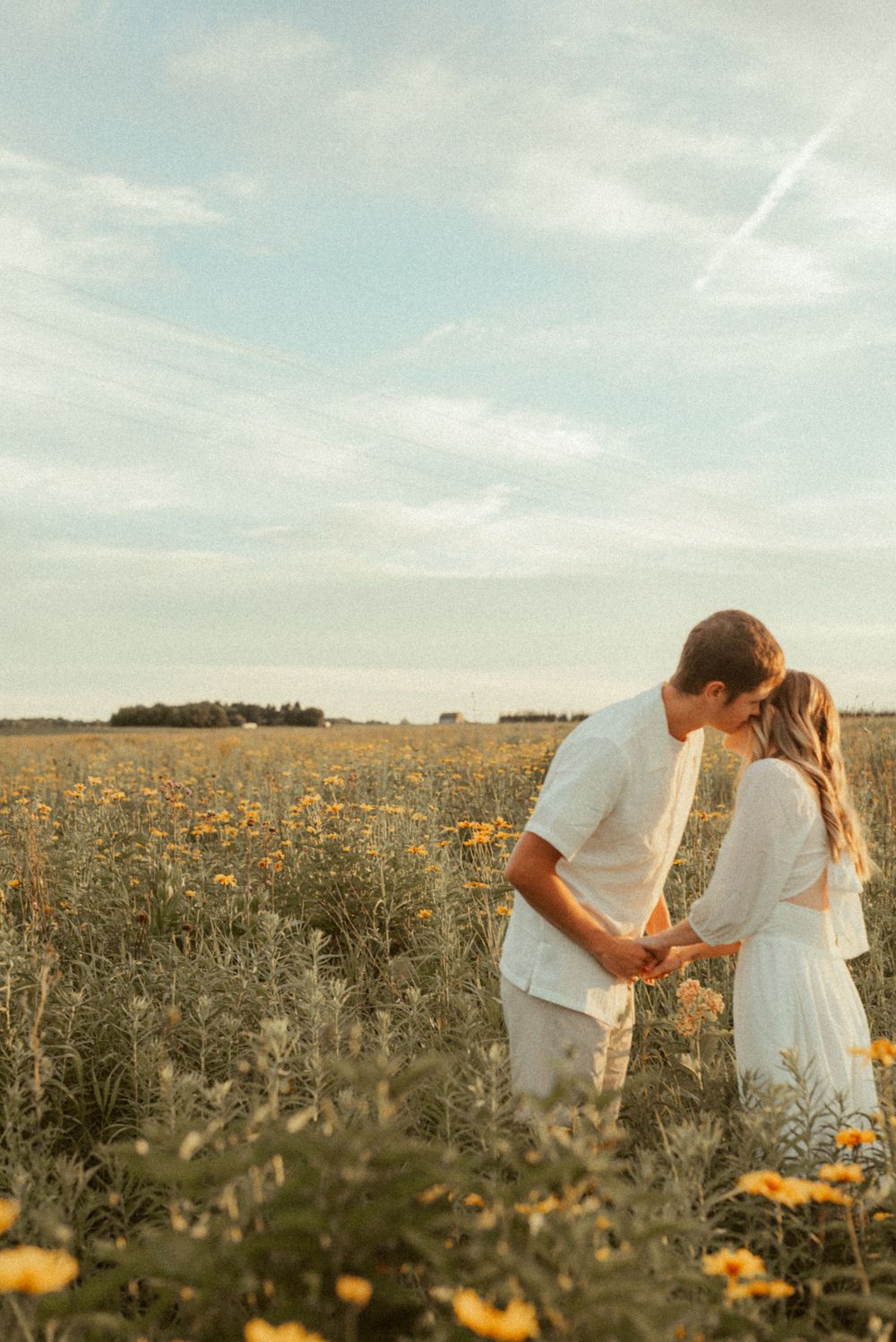 Two people in white outfits sharing tender moment in field of yellow flowers.