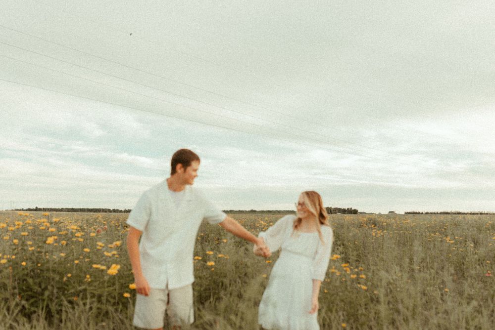 Couple holding hands while walking through field of yellow flowers at golden hour.