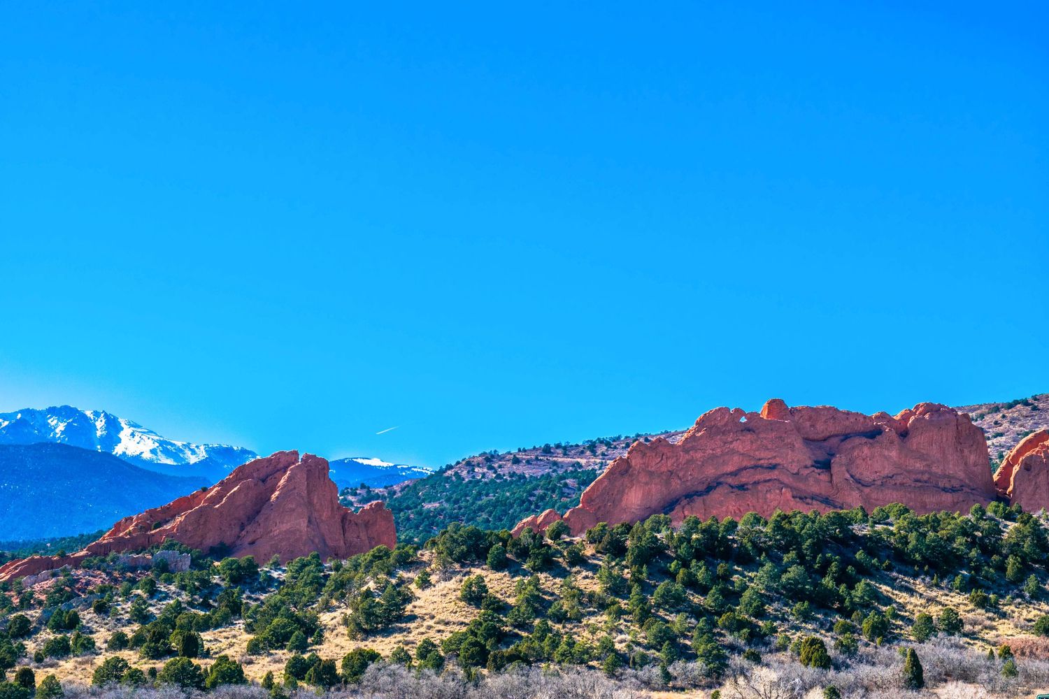 Snow-capped Pikes Peak mountain range rises behind red rock formations in Colorado landscape.