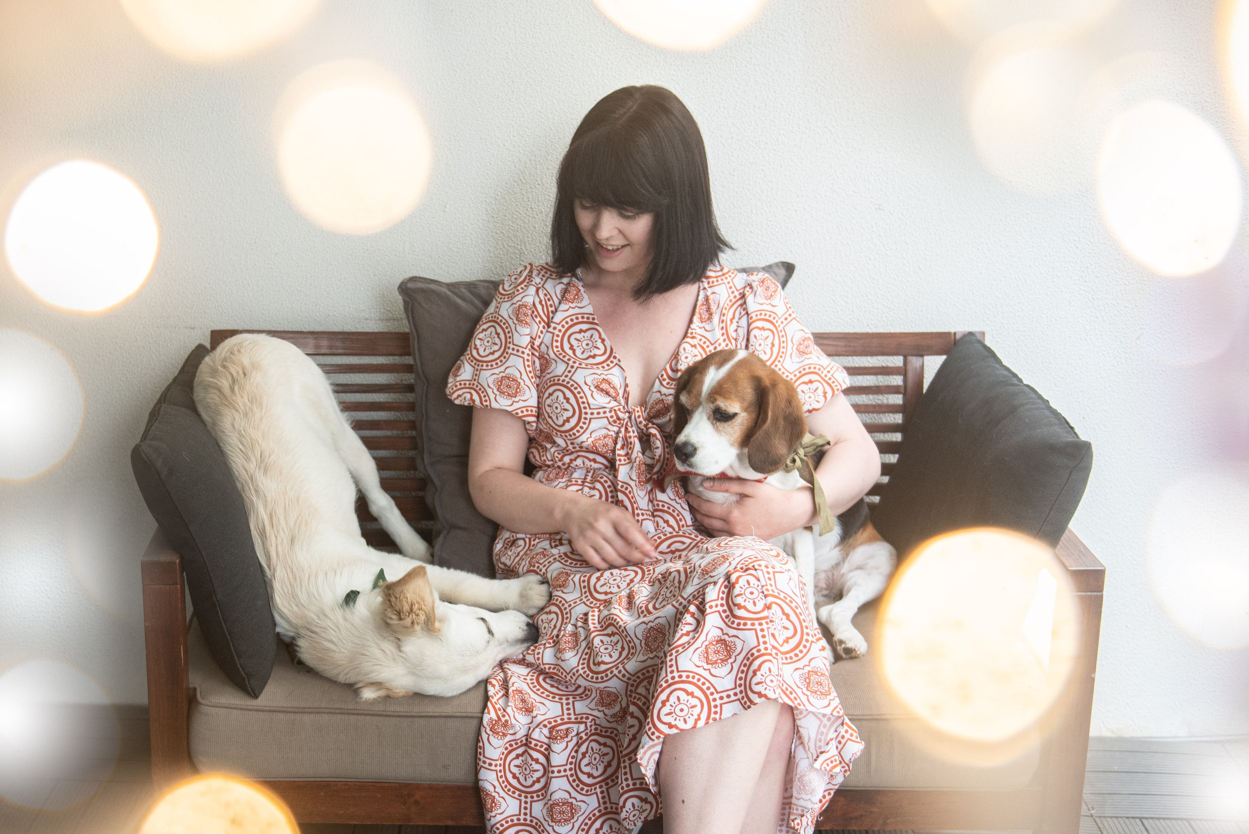 Woman in floral dress sitting on bench with two dogs in dreamy lighting.