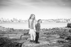 Two children standing on rocky shore with city skyline in background.