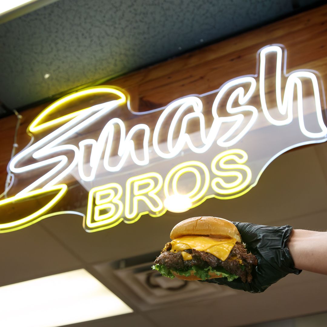 Hand holding a smash burger in front of a glowing Smash Bros neon sign.