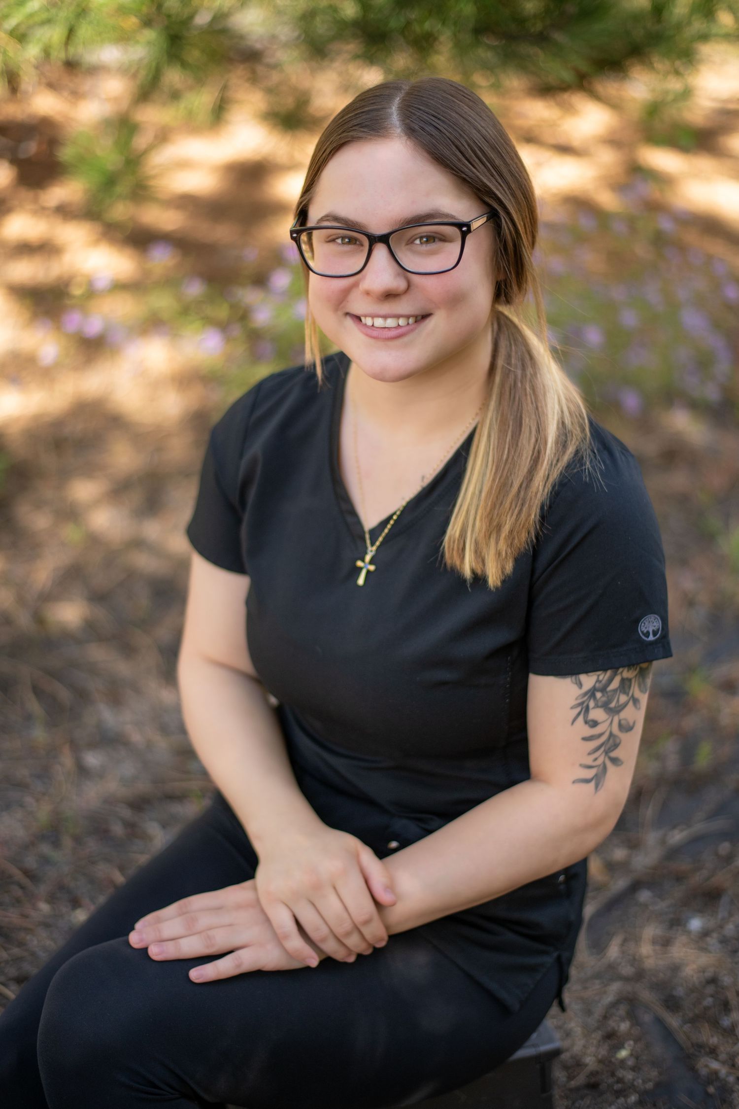 Young woman with glasses and tattoo wearing black shirt sitting outdoors.