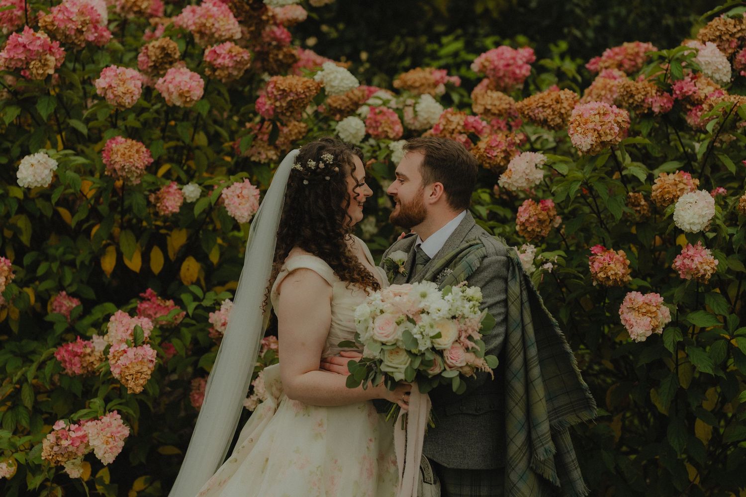 A wedding couple stands on a garden path surrounded by symmetrical topiary hedges and formal landscaping.