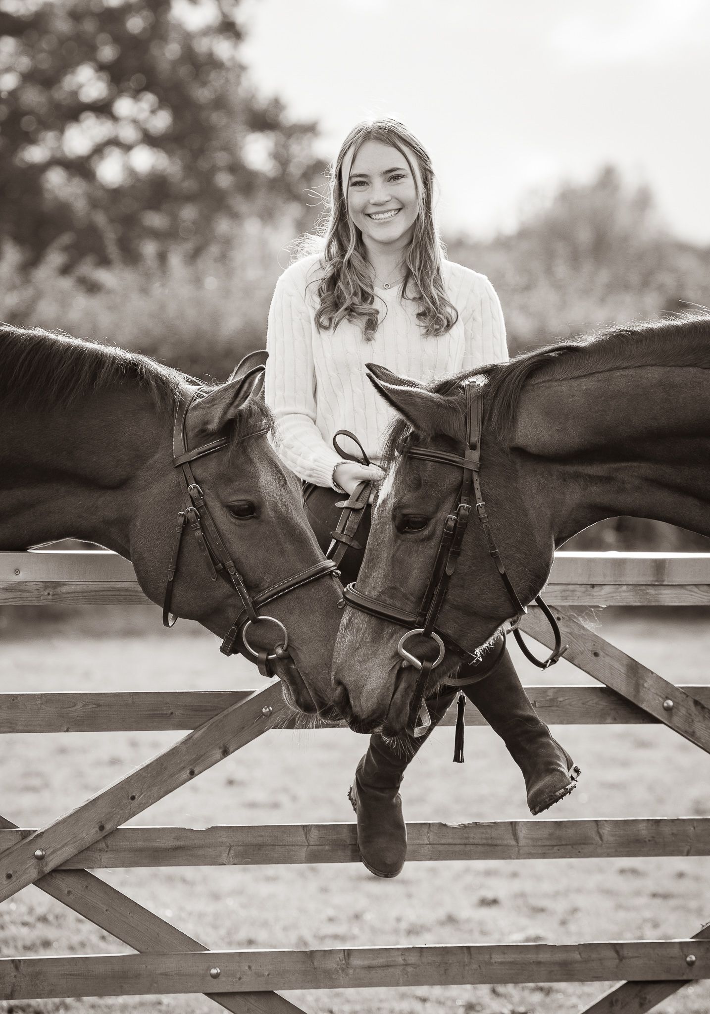 Young woman sitting on a wooden fence smiling between two horses in a black and white equestrian portrait.