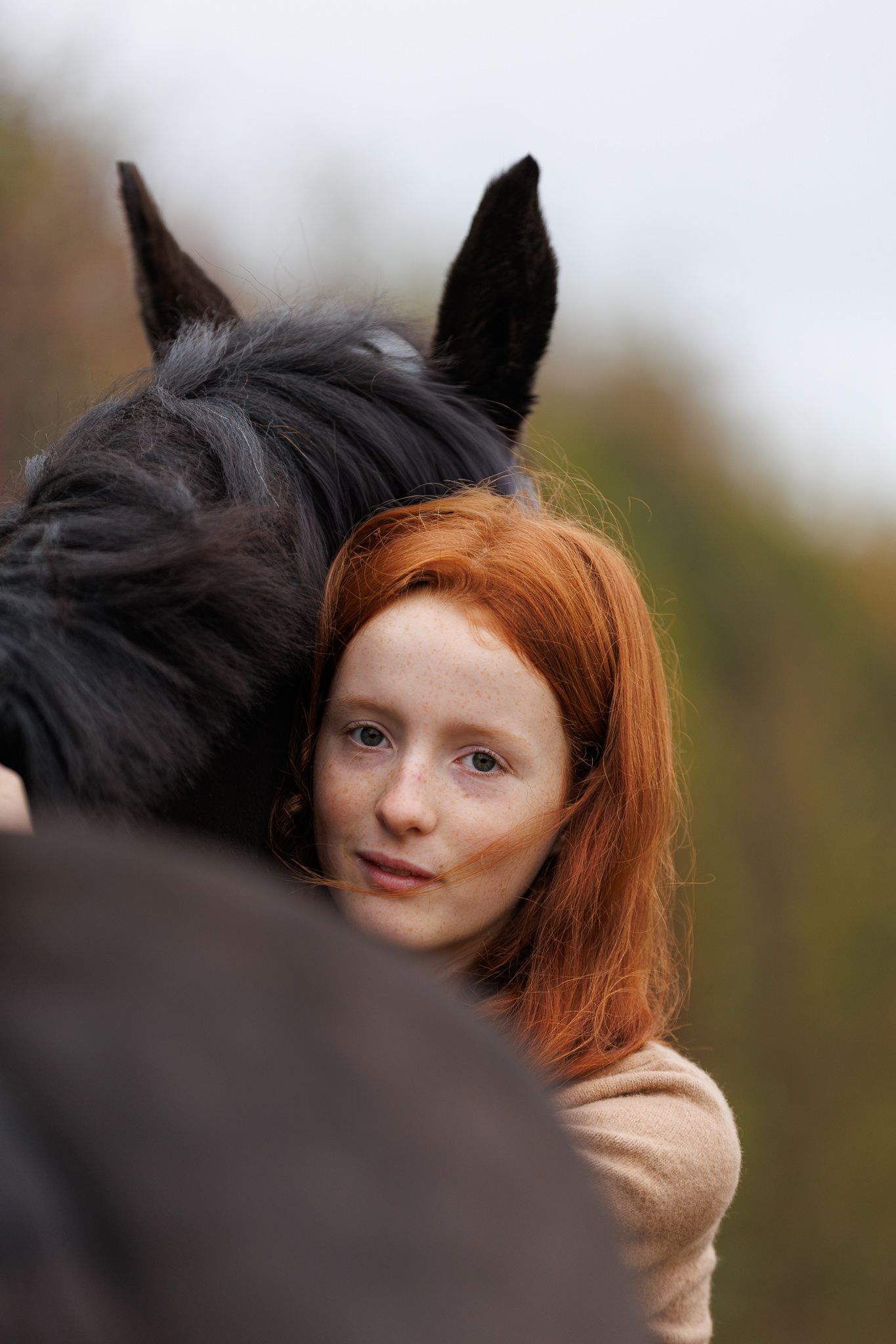 A red-haired woman tenderly rests her head against a dark horse, sharing a peaceful moment outdoors.