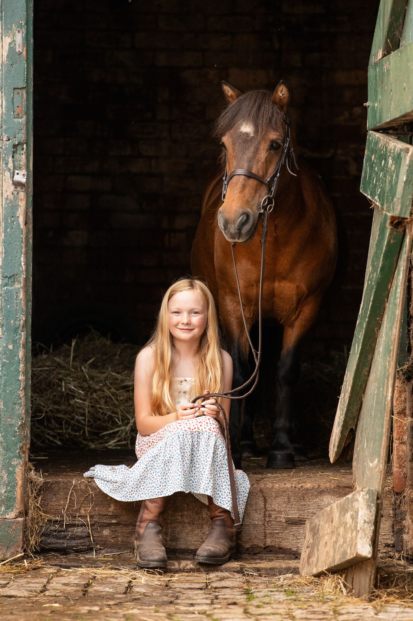 Young blonde girl in polka dot dress and boots sits smiling in stable doorway with brown bay horse behind her.