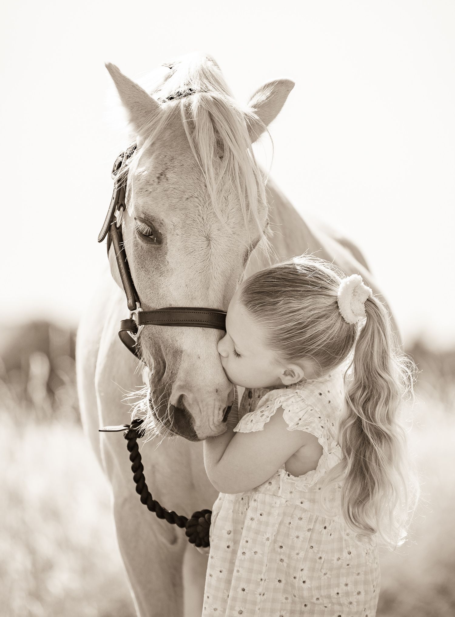 A young girl with pigtails tenderly kisses a white horse on the nose in a soft sepia-toned outdoor photo.