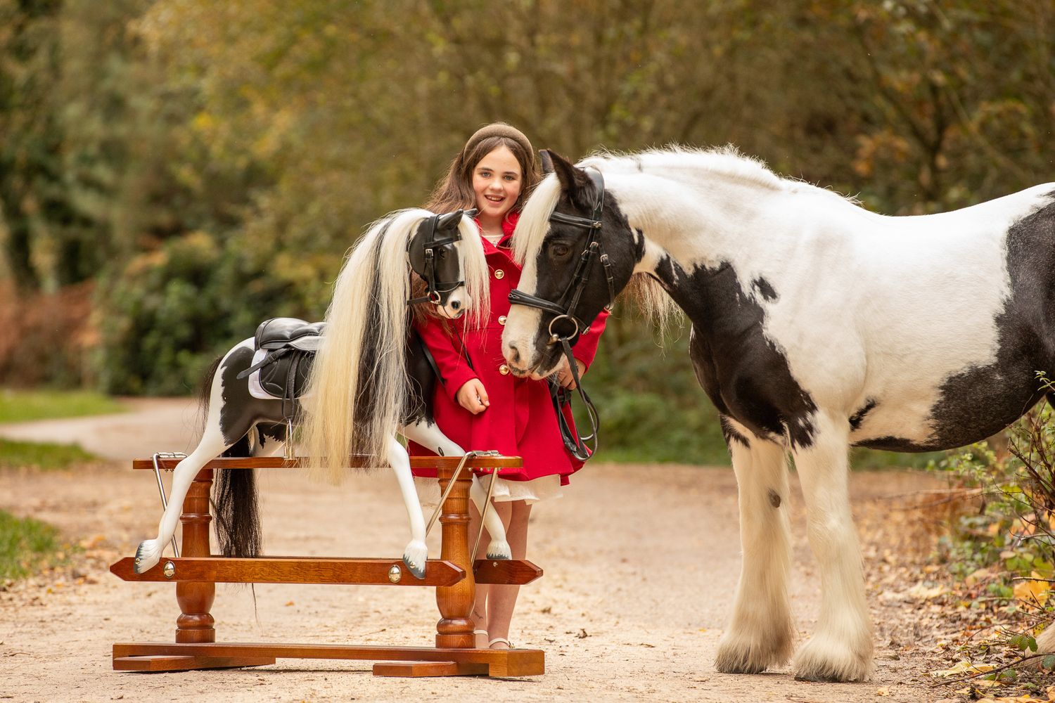 Girl in red dress stands between a black-and-white Gypsy Vanner horse and a wooden rocking horse outdoors.