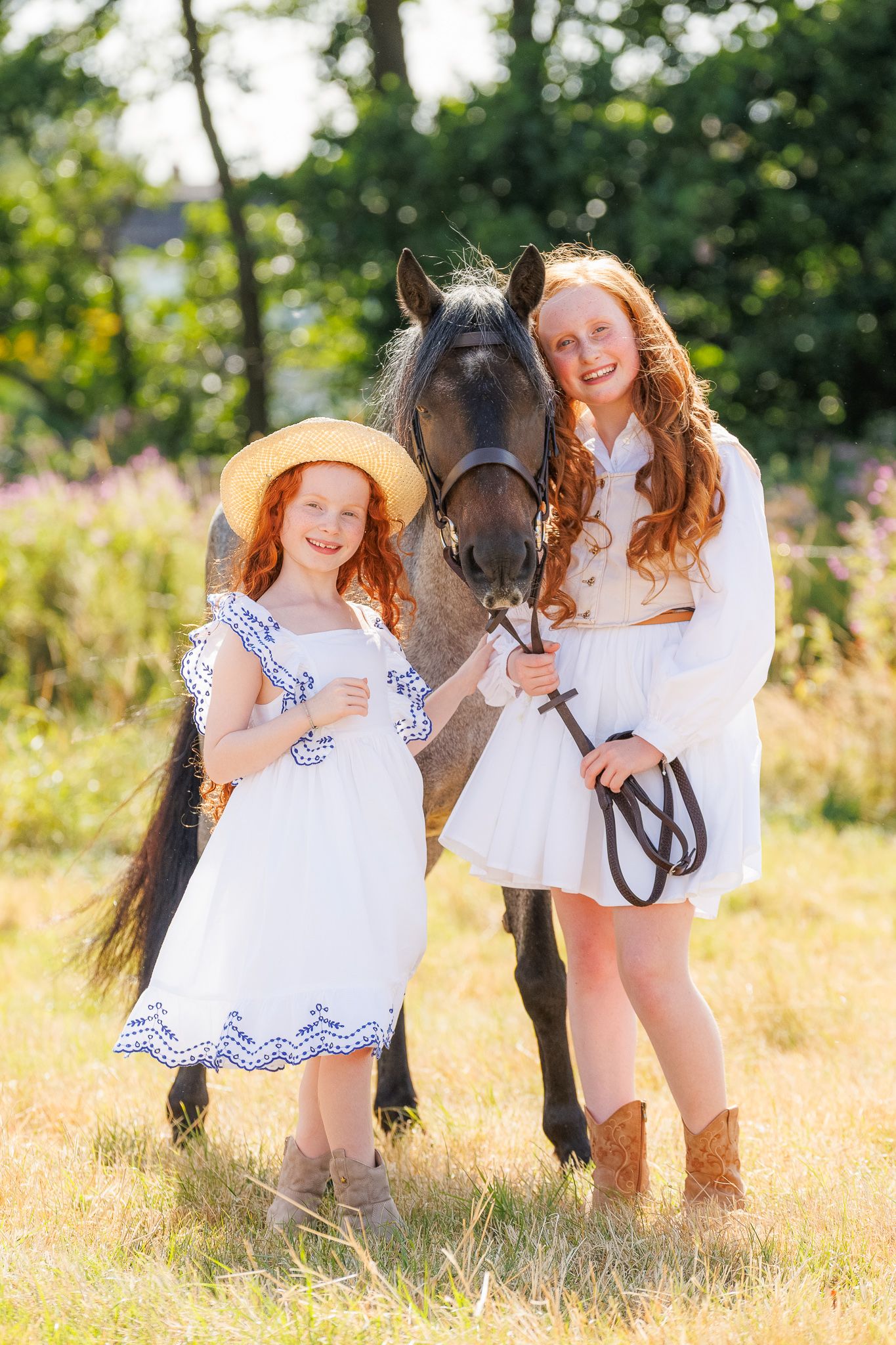 Two redhead girls in white dresses pose with a dark horse in a sunny green meadow.