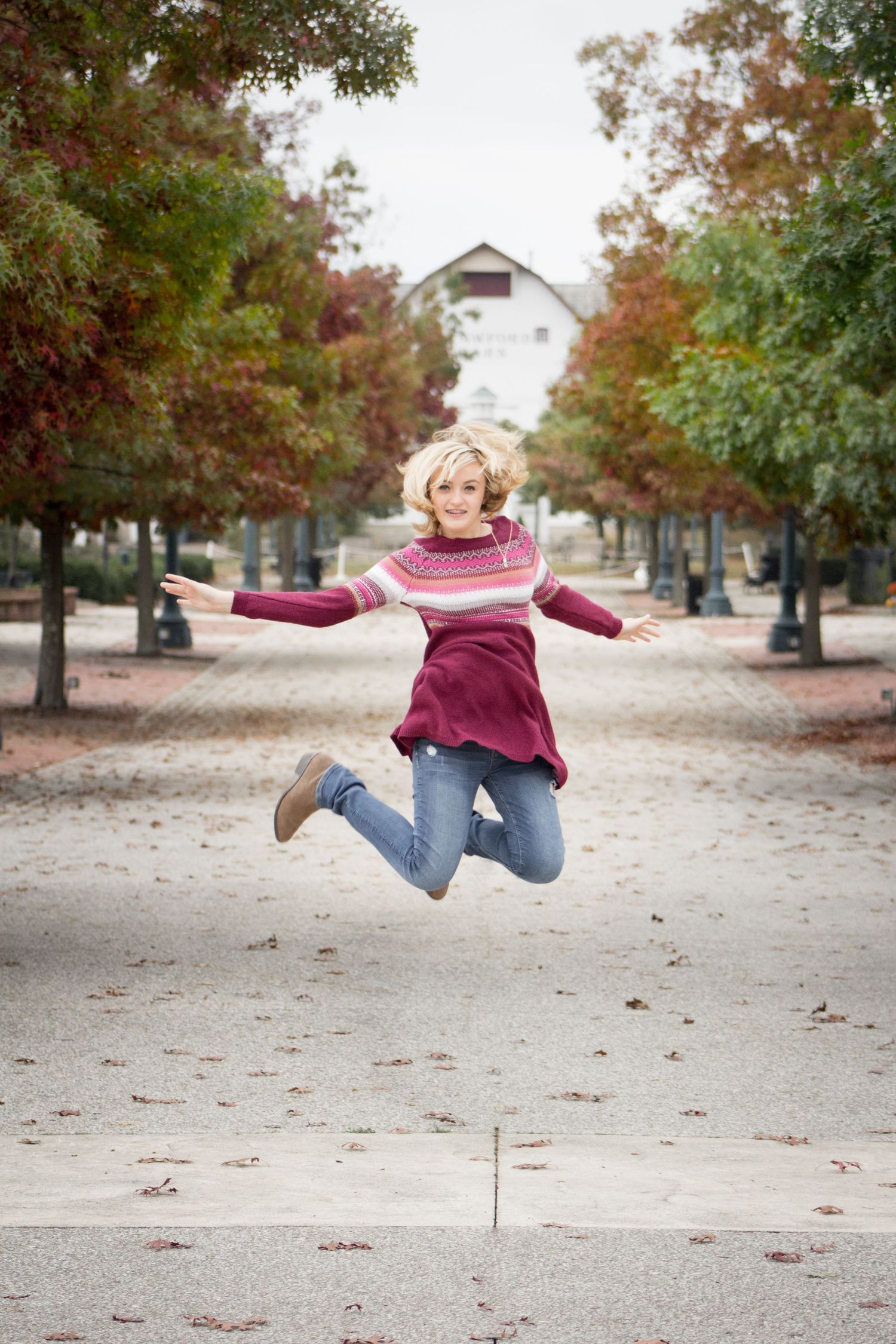 Joyful blonde woman jumping in tree-lined pathway during autumn season.