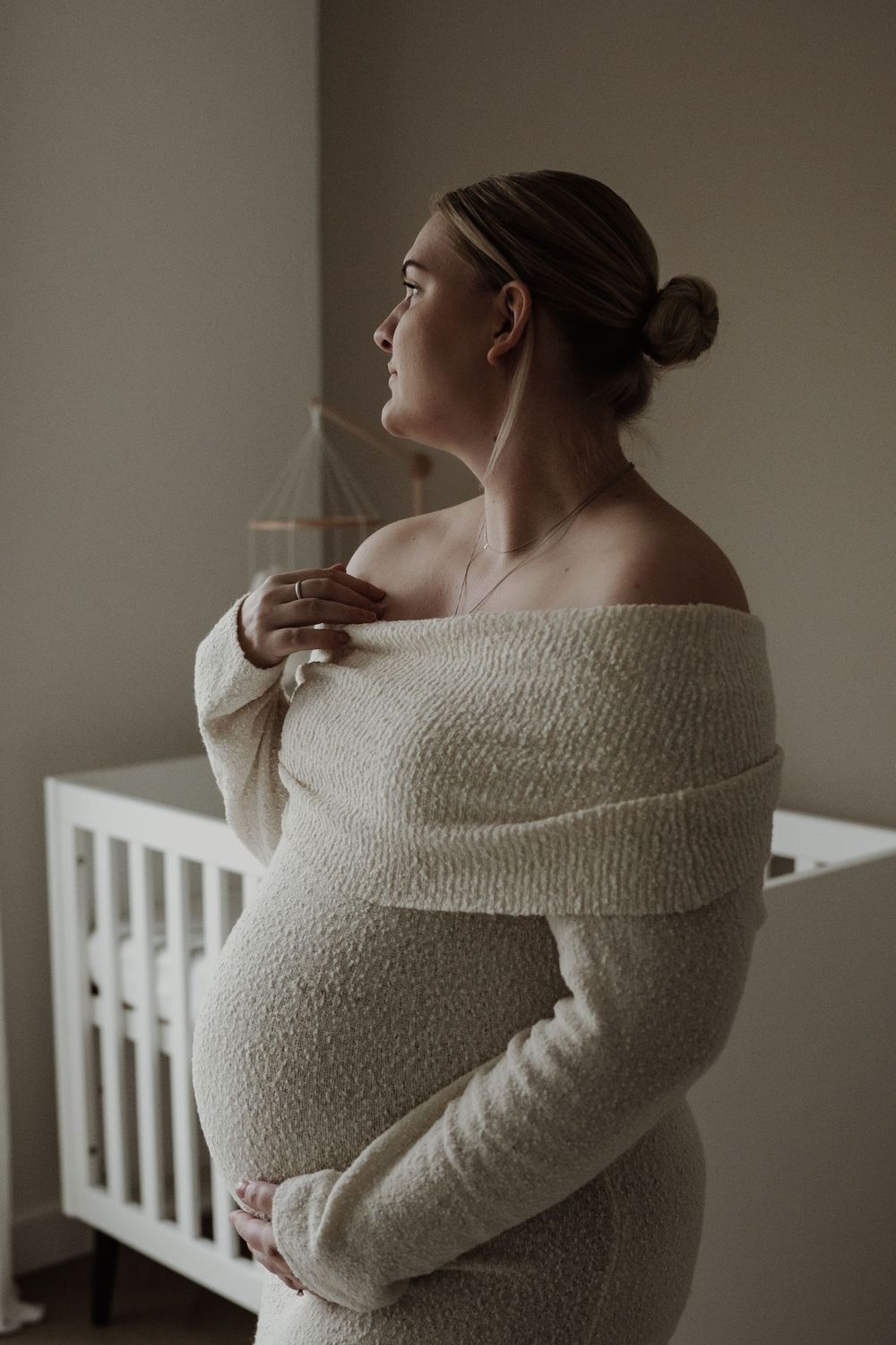 Side profile of an expectant mother in a cozy white sweater standing by a crib in a peaceful nursery setting.