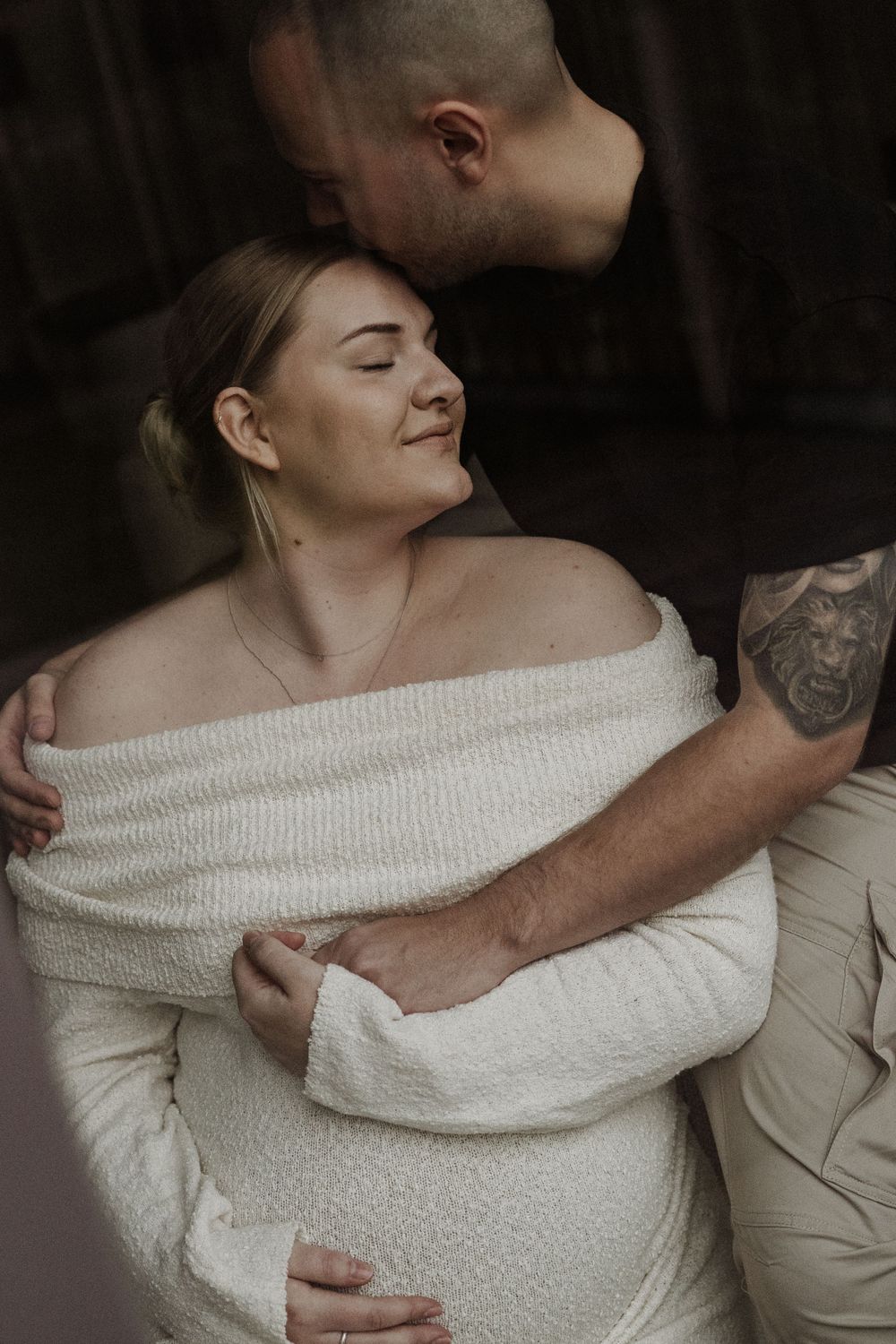 Romantic couple embraces intimately as woman in off-shoulder white sweater smiles with eyes closed in moody lighting.