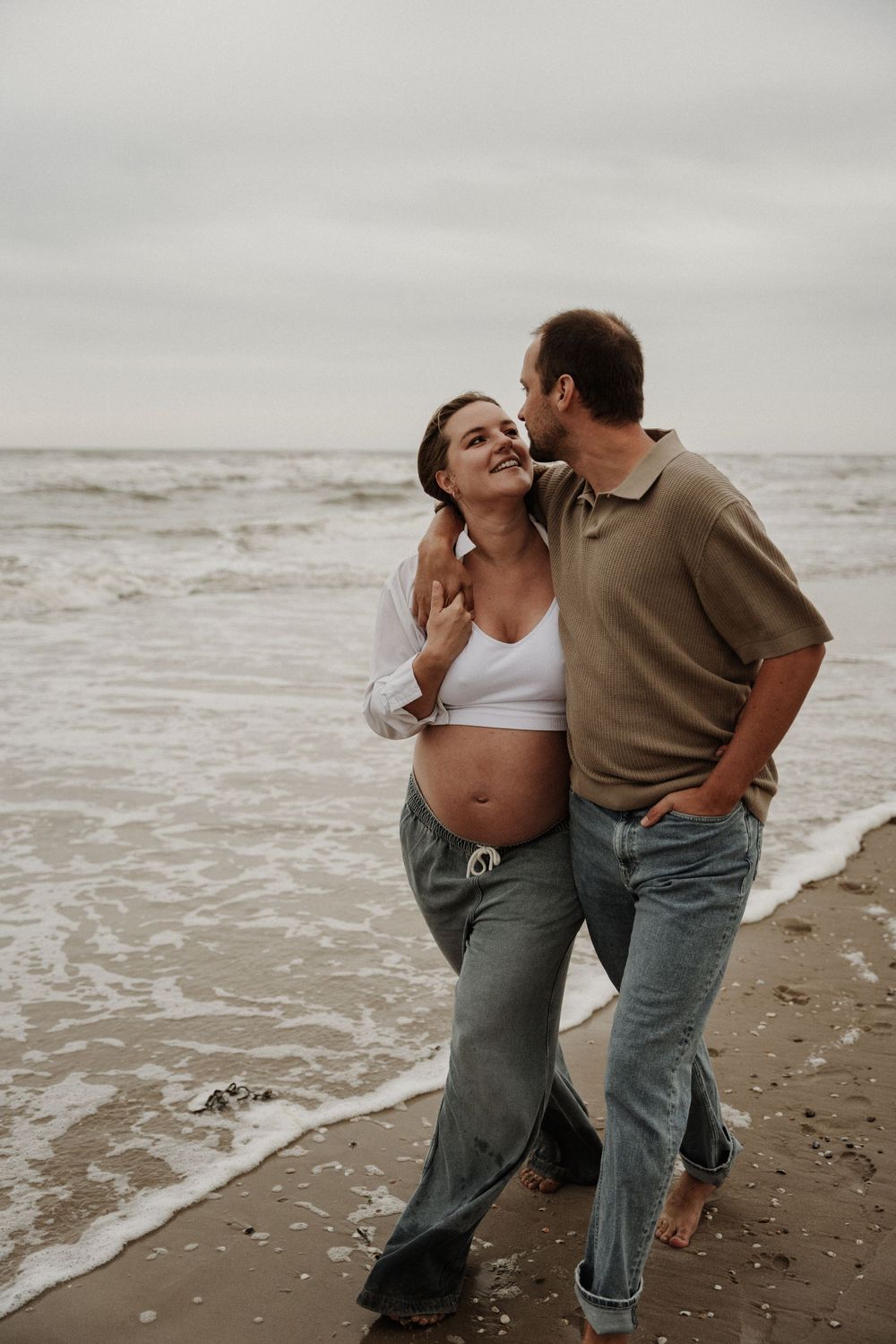A couple shares a joyful moment together on a cloudy beach during a maternity photoshoot.
