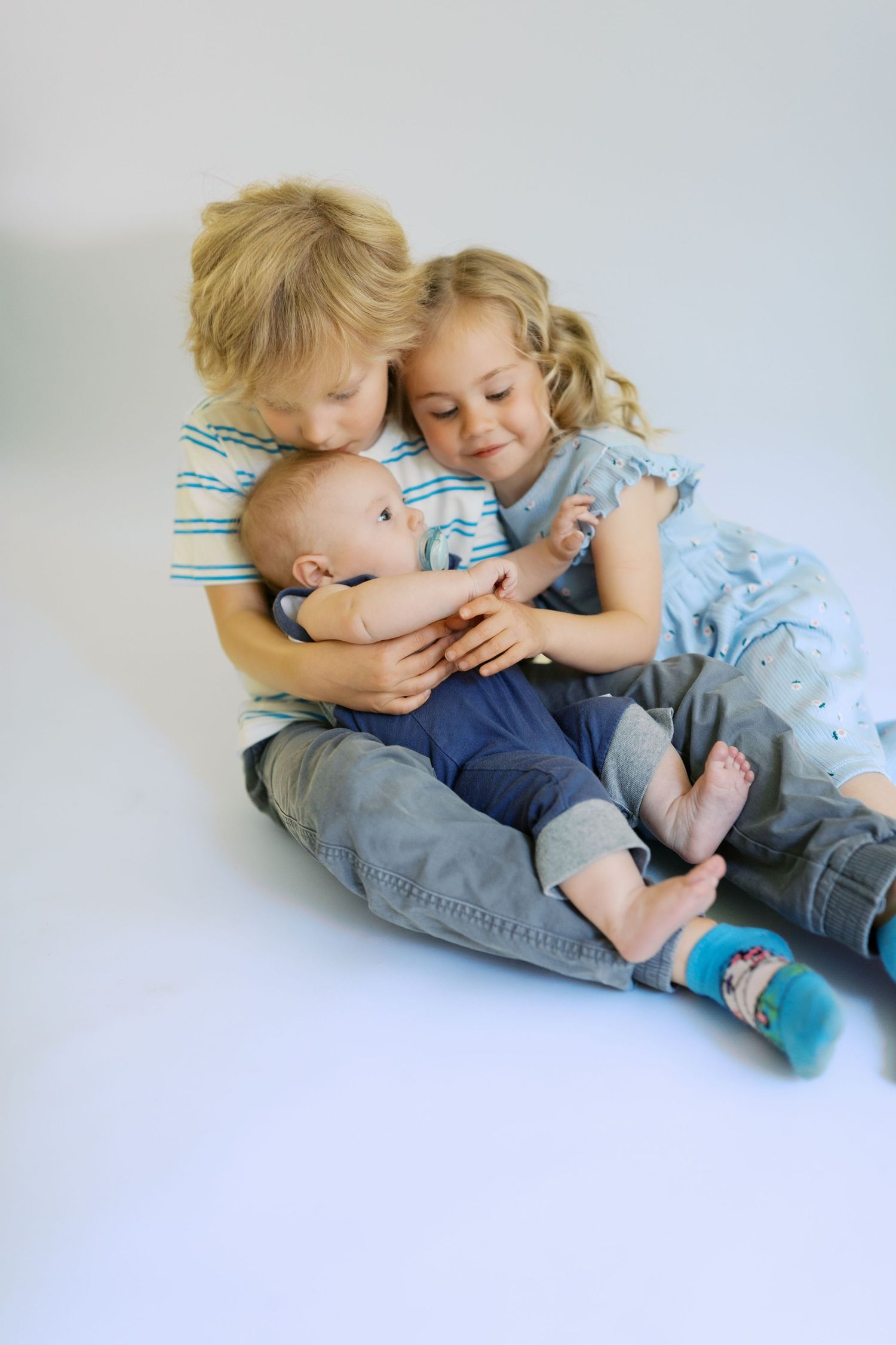 Two blonde siblings lovingly cuddle a baby on a white background in a sweet family portrait.