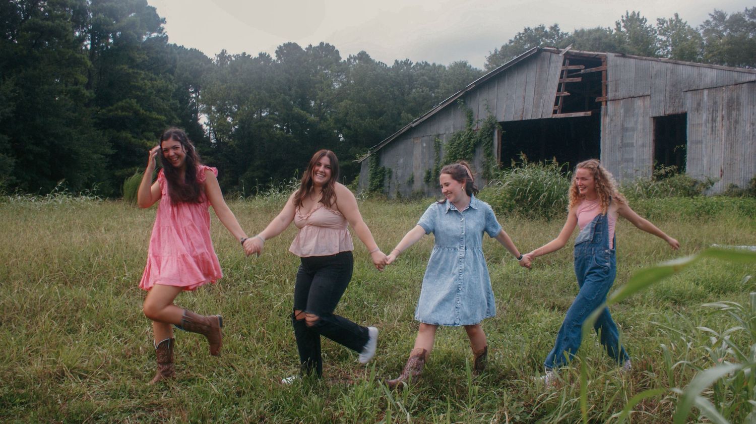Four friends in casual outfits dancing joyfully in a field near an old barn.