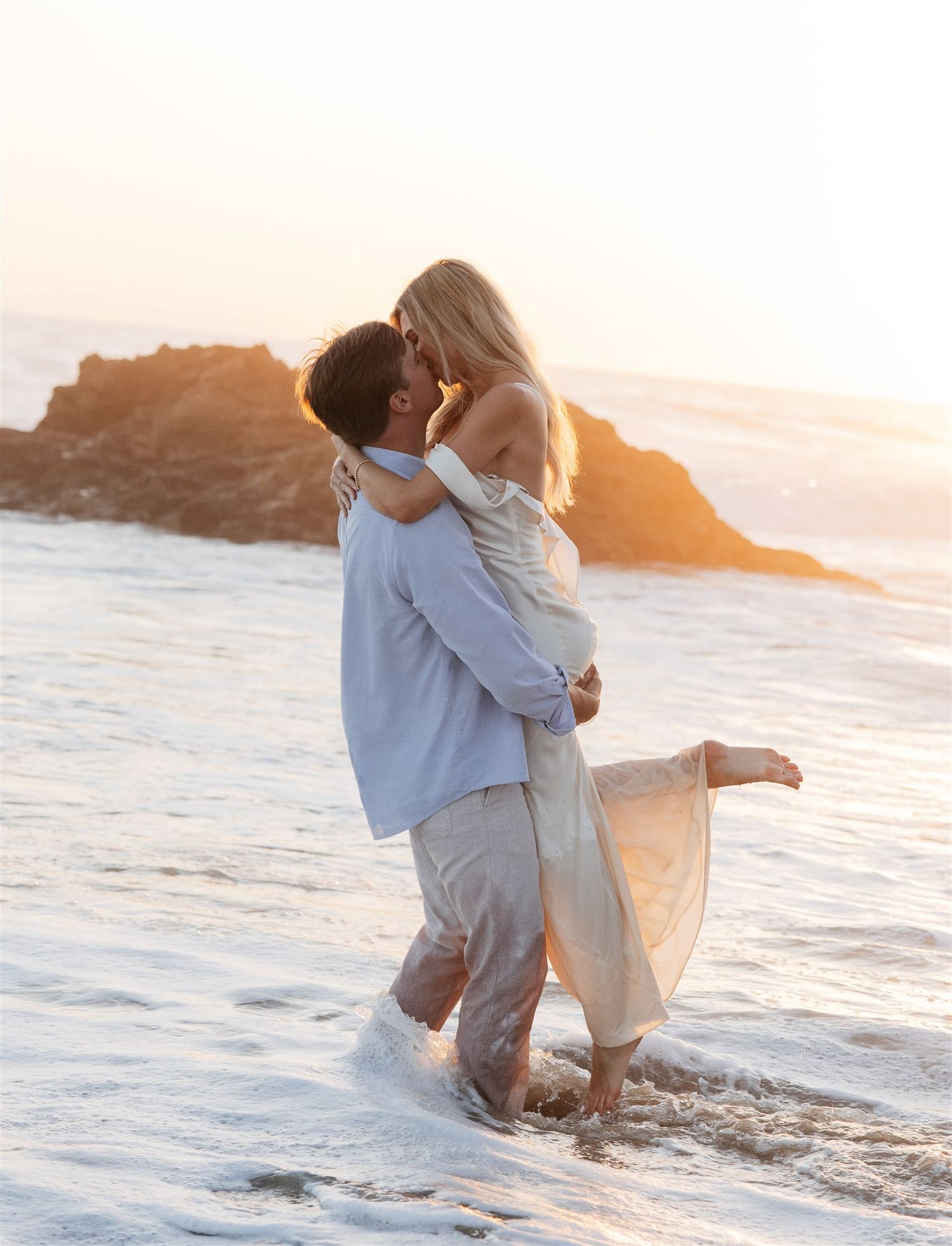 A couple embraces and kisses on a sandy beach at dusk with ocean waves in the background.