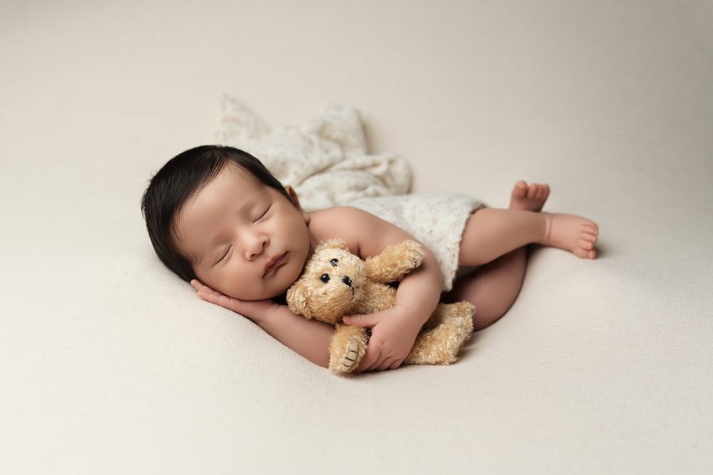 Sleeping baby cuddled with small teddy bear on white blanket in peaceful newborn photography session.