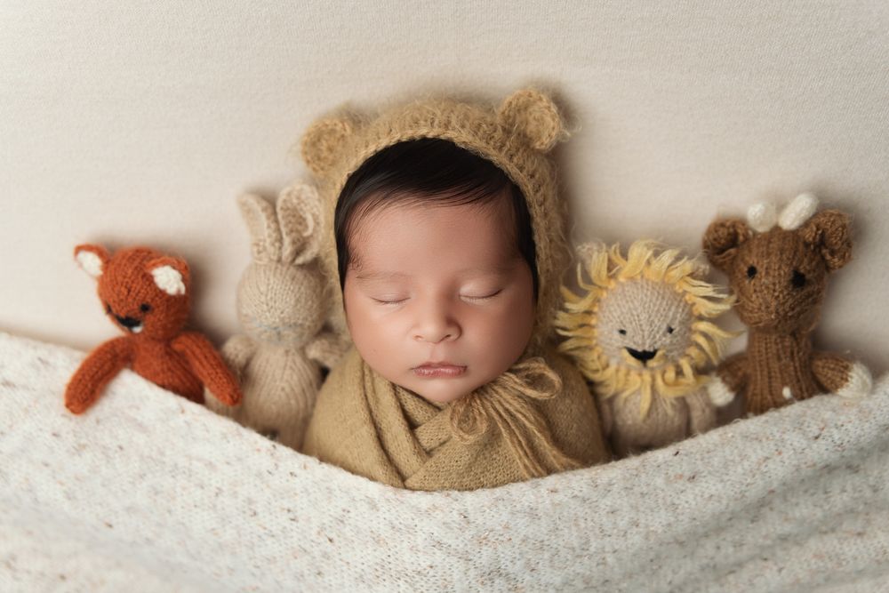 Sleeping newborn in brown outfit surrounded by plush stuffed animals on white textured blanket.