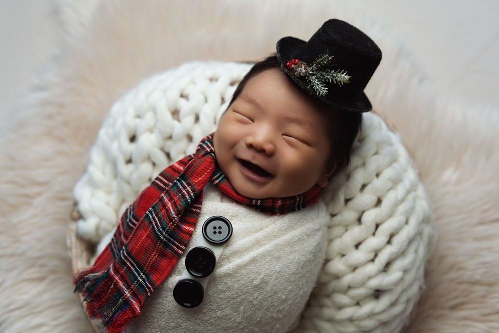 Newborn baby wearing black top hat and plaid scarf sleeps peacefully on chunky knit blanket.