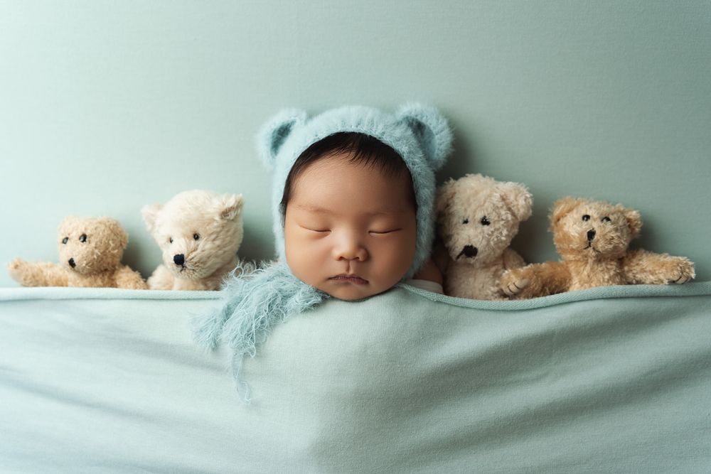 Sleeping newborn baby surrounded by plush teddy bears on a soft mint green blanket.
