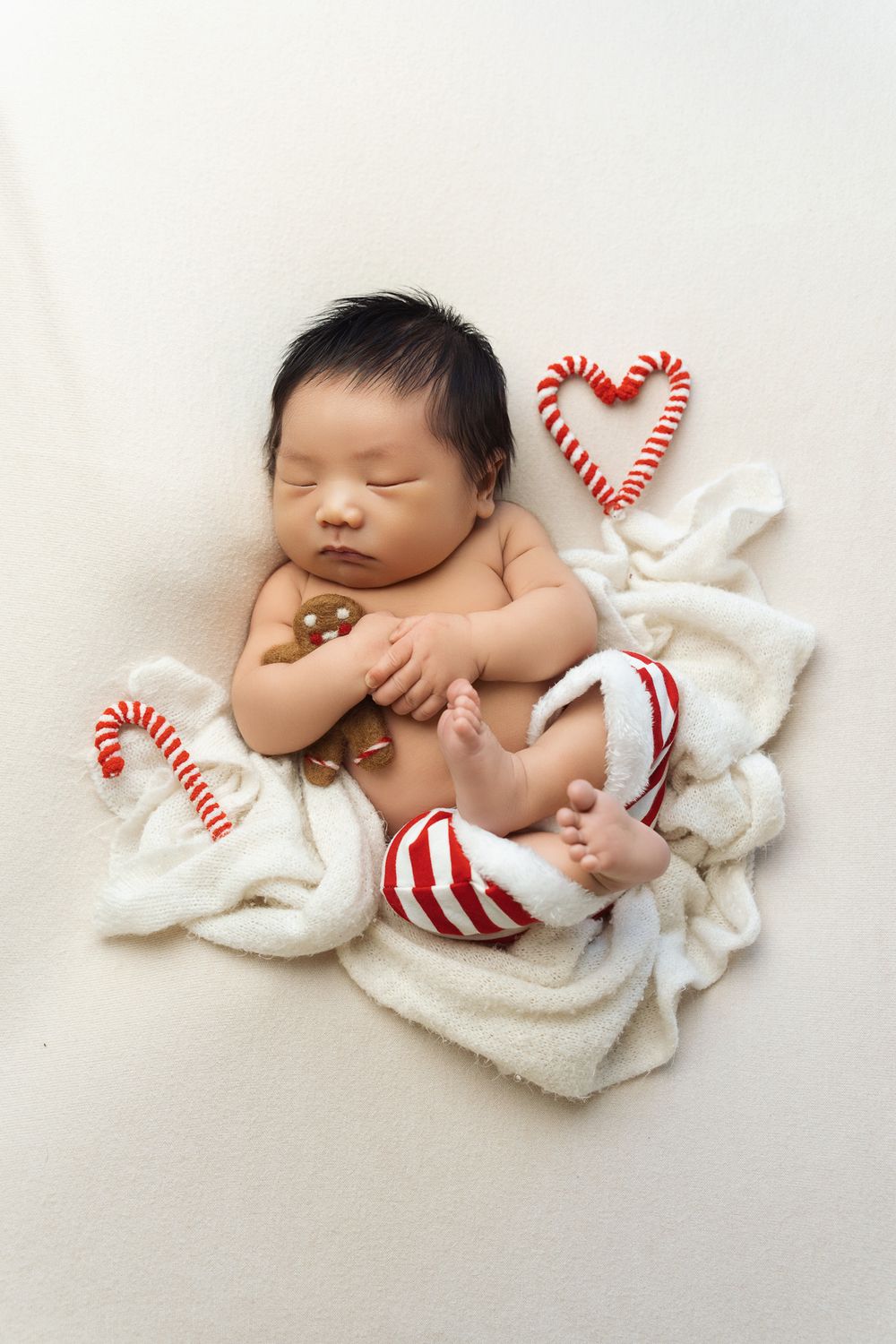 Sleeping newborn baby swaddled in white blanket with red and white candy cane heart decorations for Christmas photo.