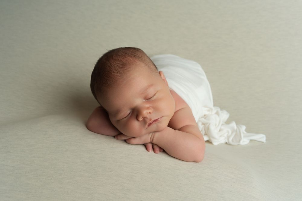 A sleeping newborn baby wrapped in white fabric lies peacefully against a soft neutral background.