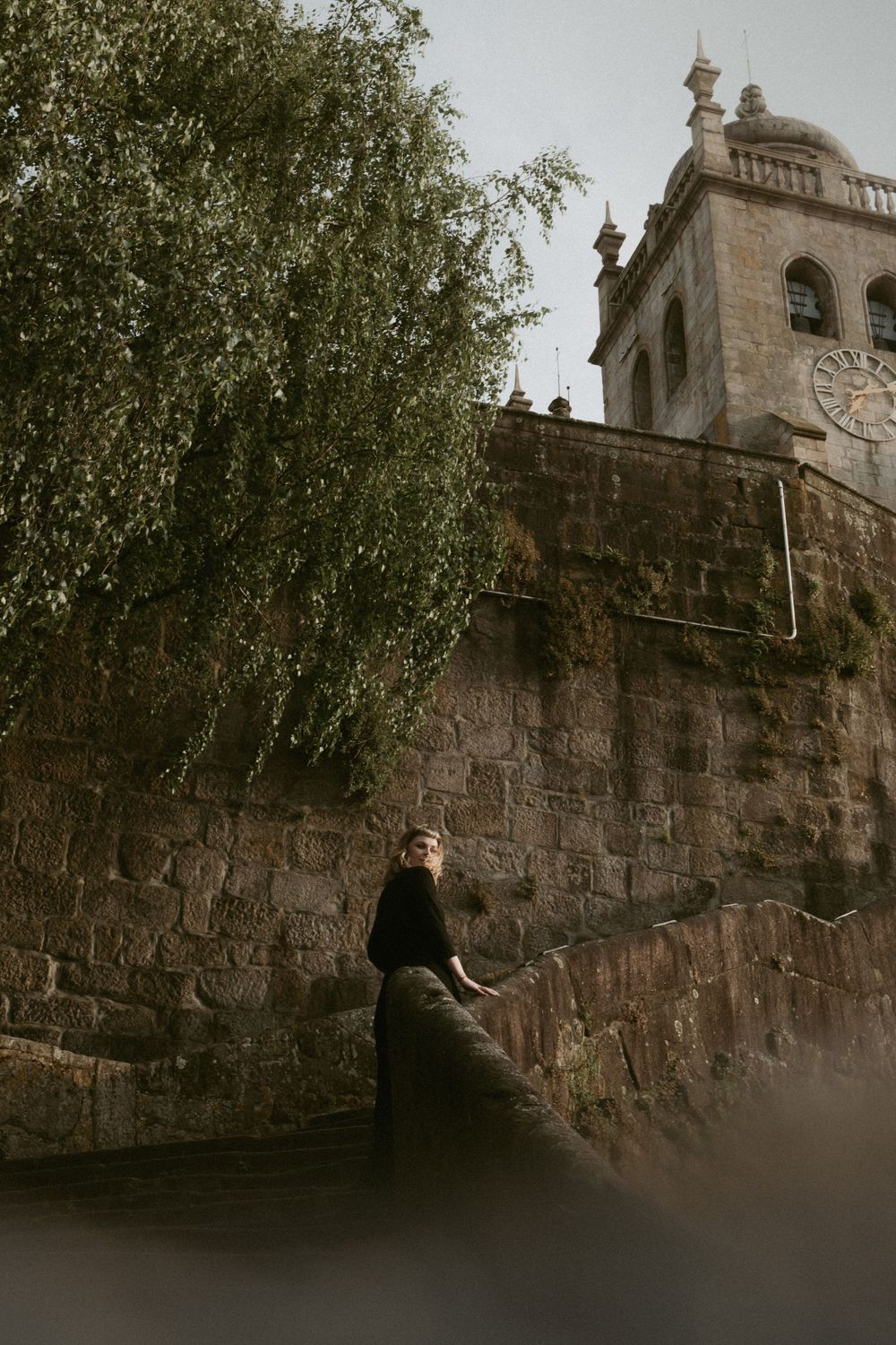 Person in black dress sitting on stone wall near historic church tower.