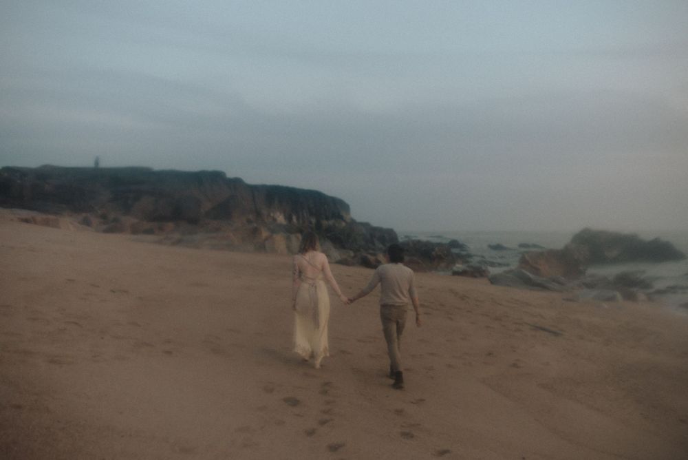 Two people walking hand in hand along a misty coastal beach with cliffs in the background.
