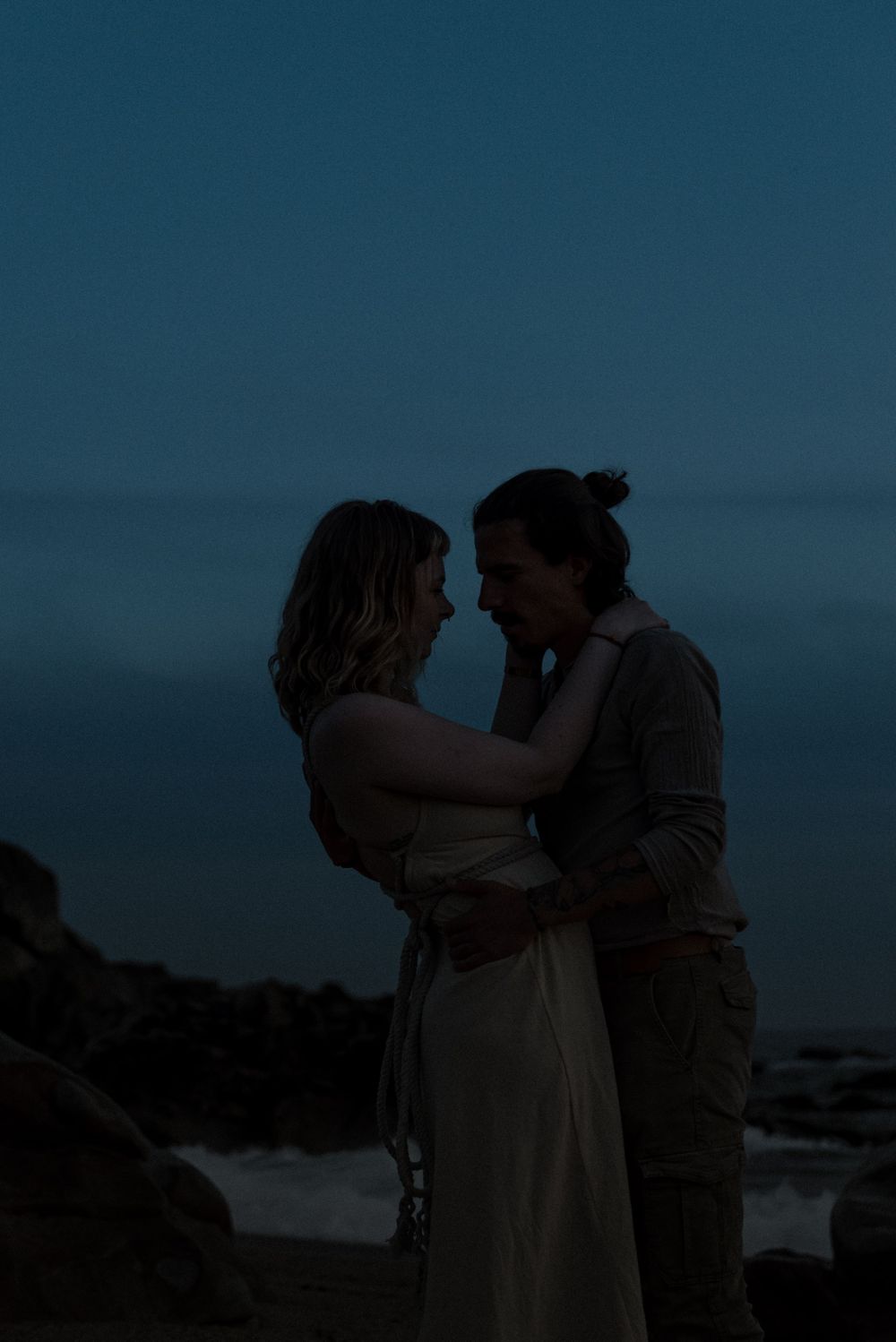A romantic couple embraces on a rocky beach at dusk with the ocean as their backdrop.