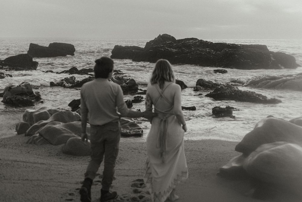 Black and white photo of a couple walking along a rocky coastline overlooking the ocean.