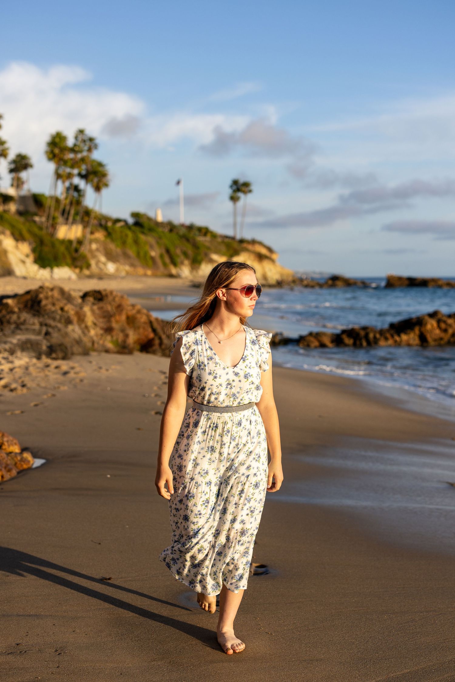 Person in floral dress walking along scenic beach at sunset with cliffs and ocean in background.