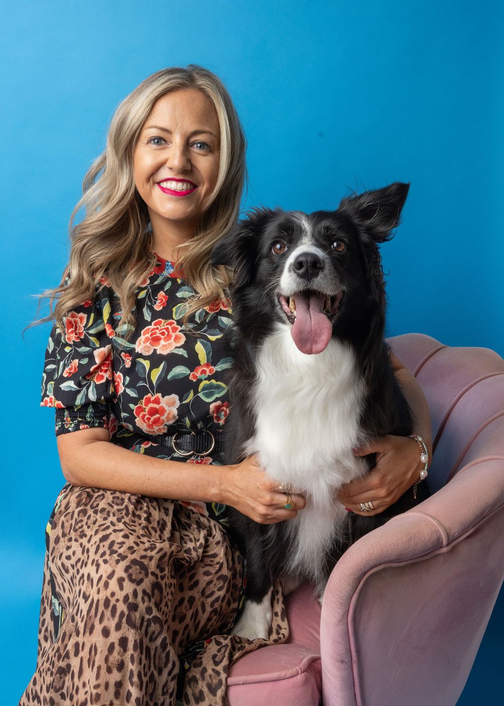 Smiling blonde woman in floral top and leopard print pants posing with a black and white Border Collie on a pink chair.