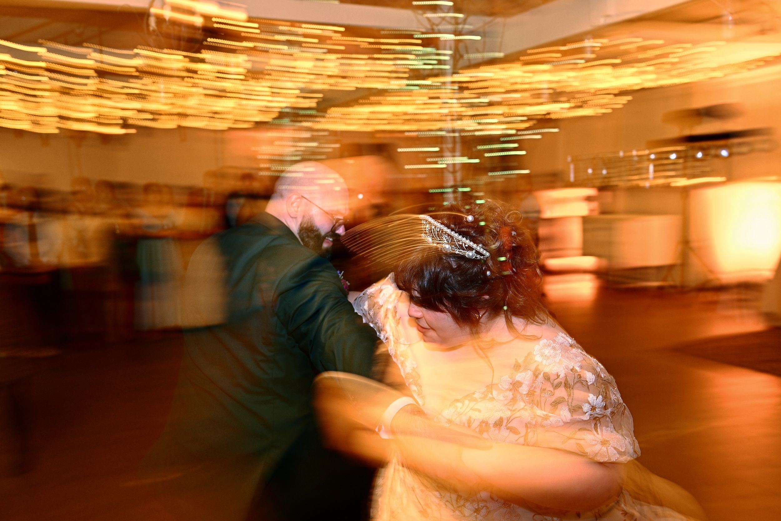 Blurred motion shot of bride and groom sharing first dance at wedding reception with warm golden lights.