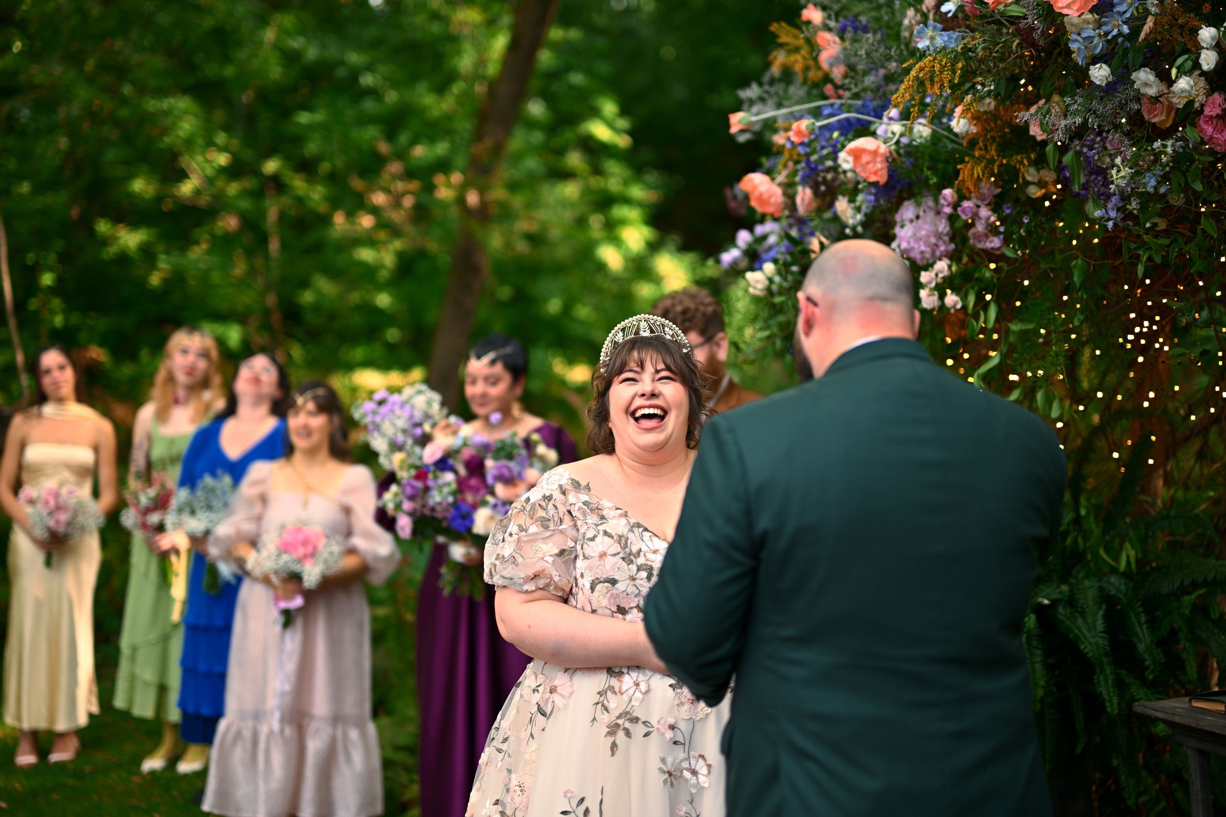 Bride laughing joyfully at outdoor wedding ceremony with bridesmaids in colorful dresses behind her.