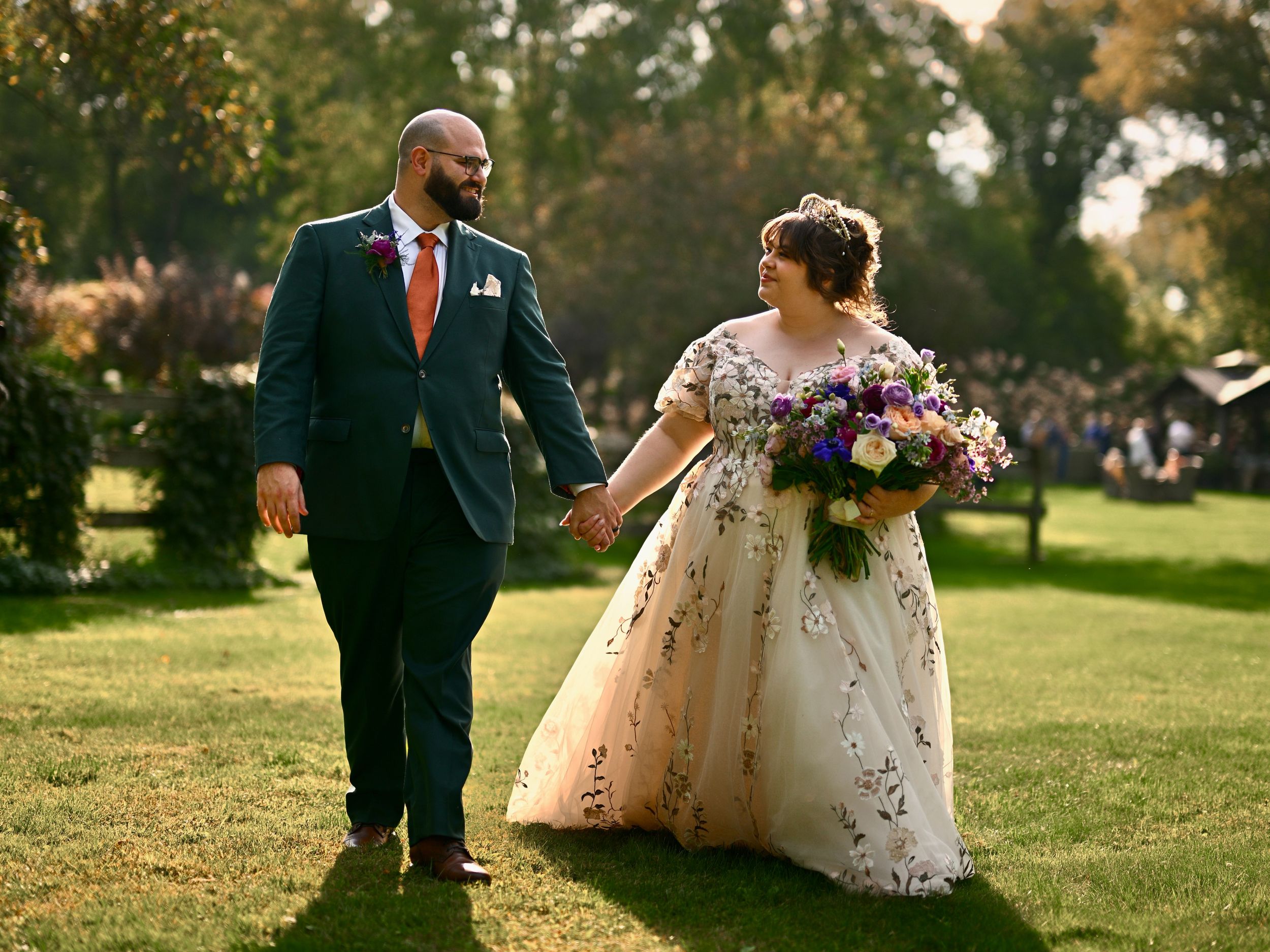 Bride and groom walking hand in hand on green lawn, bride holding colorful floral bouquet.