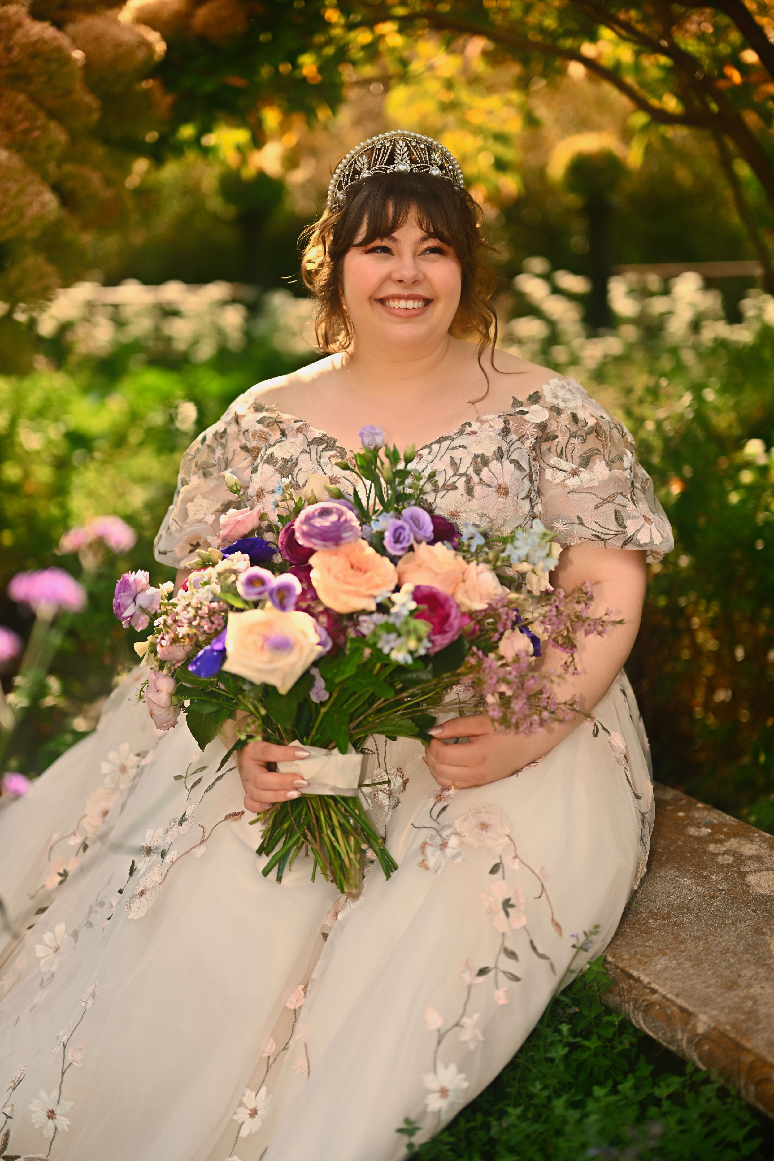 Smiling bride in floral embroidered gown holding vibrant purple and pink bouquet in garden setting.