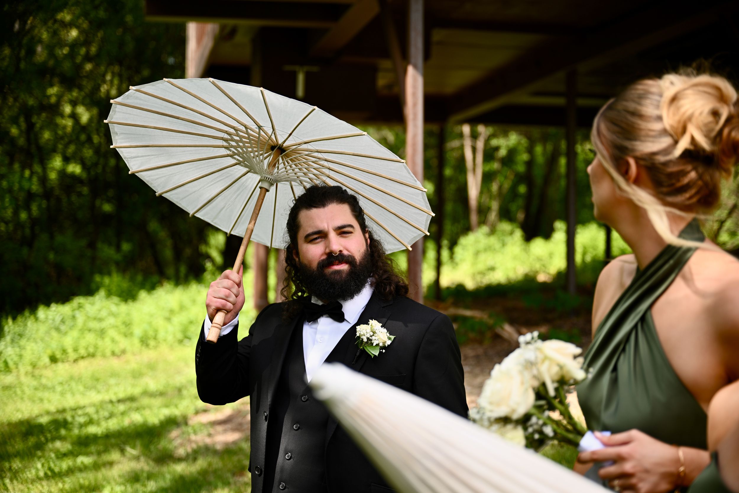 Bearded groom in tuxedo holds white parasol umbrella at outdoor wedding ceremony with bridesmaid.