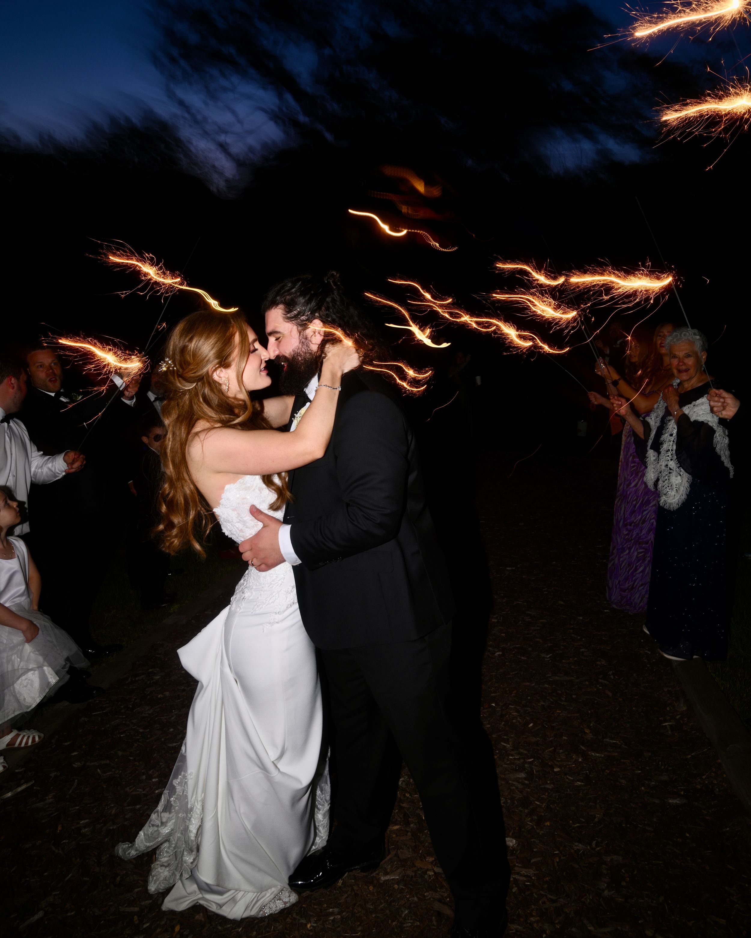 Bride and groom share romantic kiss surrounded by sparklers at night wedding sendoff celebration.