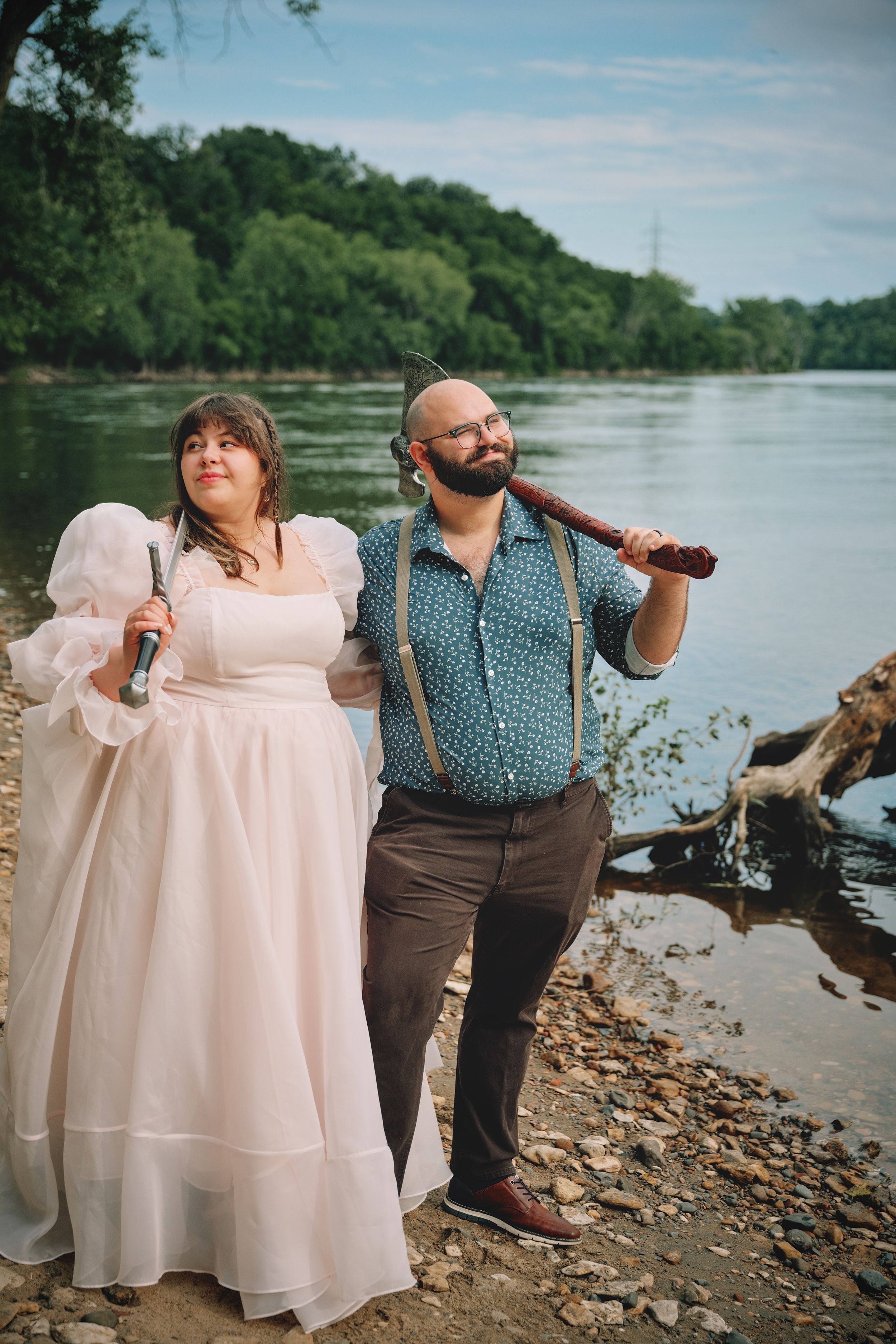 Couple posing by riverside holding swords, woman in white dress and man in suspenders.