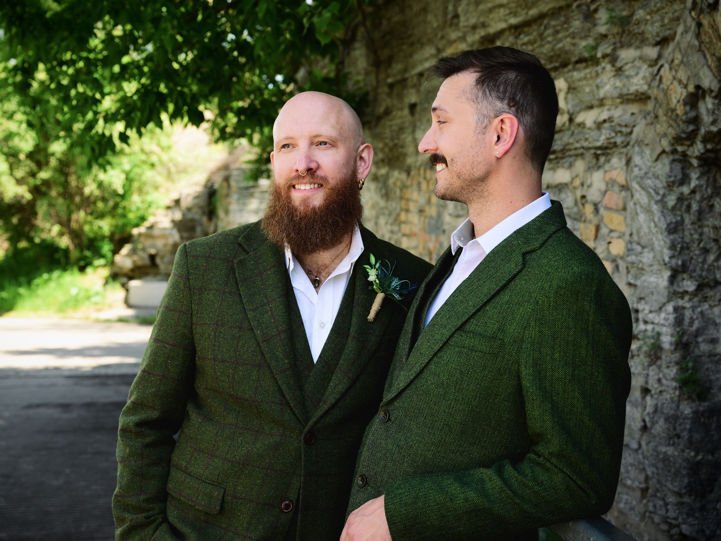 Two grooms in matching green tweed suits smiling together outdoors near a stone wall on their wedding day.
