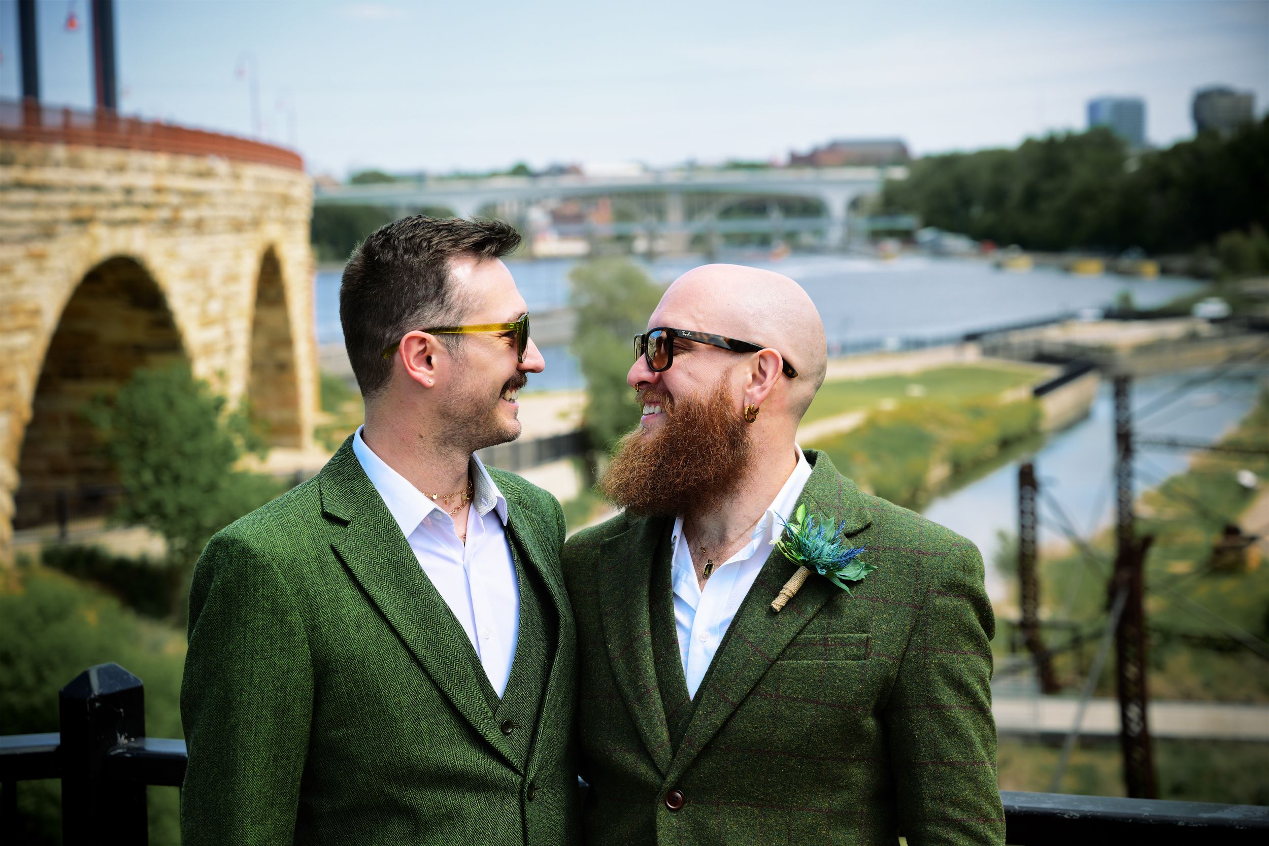 Two grooms in matching green suits and sunglasses share a romantic moment outdoors near a scenic river bridge.