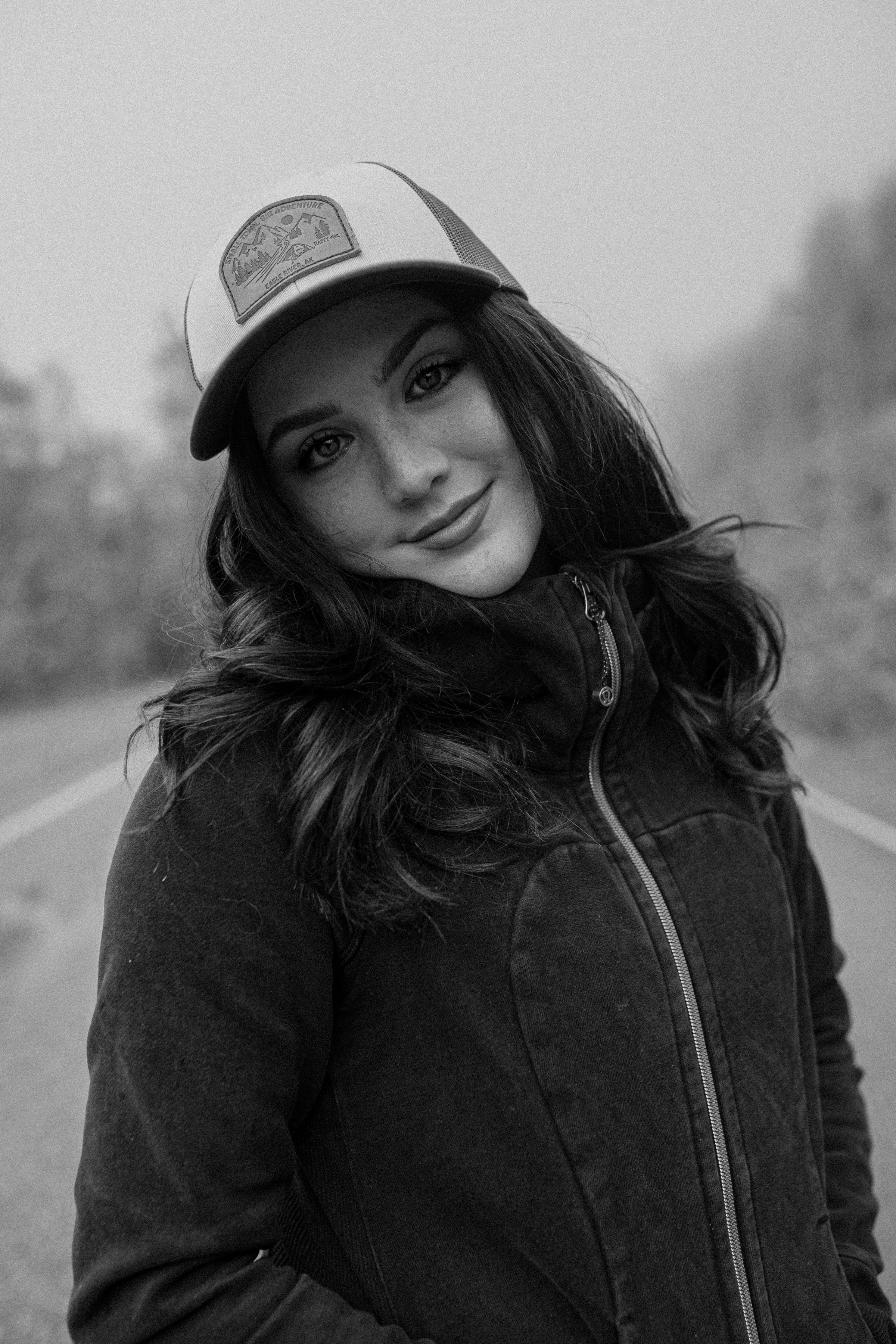 Black and white portrait of a girl wearing a trucker hat and winter jacket on a foggy road.