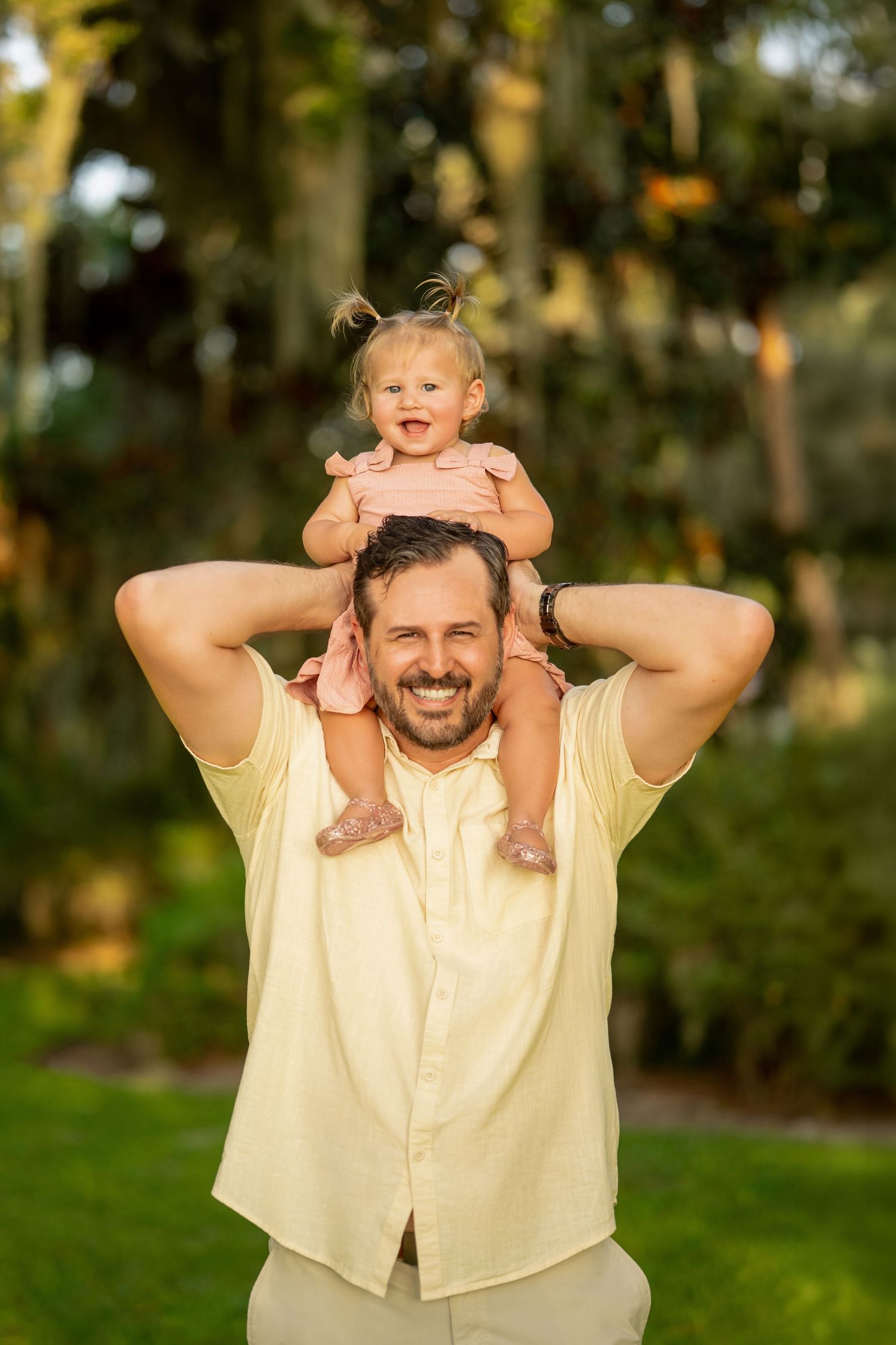 Smiling father holds laughing baby girl on his shoulders outdoors in a lush green tropical garden setting.