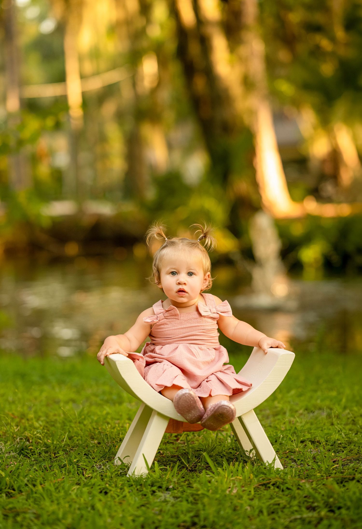 Adorable baby girl in pink dress sitting on small white chair on green grass with tropical garden background.